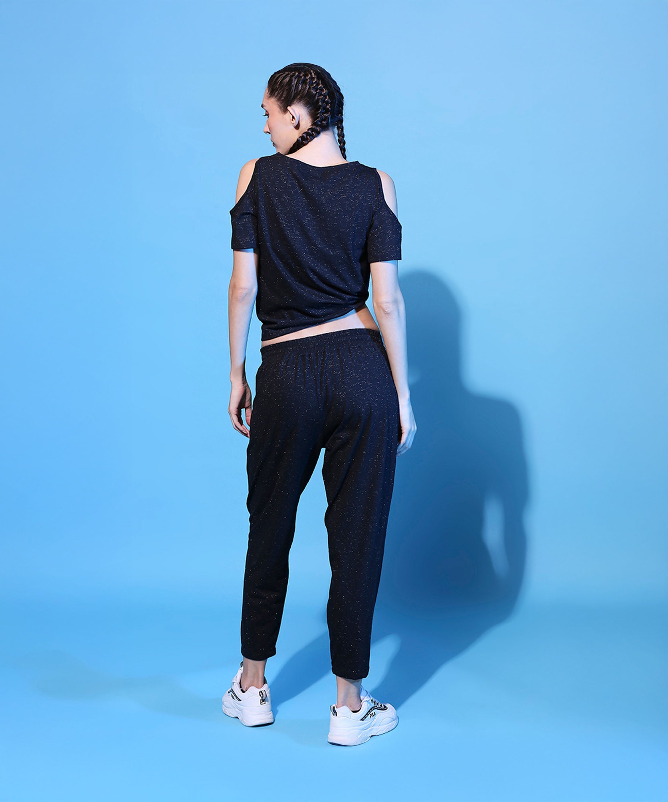Woman with braided hair wears the Shimmer Cold Shoulder Tee (Customised) and Side Tie Up Shimmer Yoga Pants - AW21, standing on a blue background, facing away.