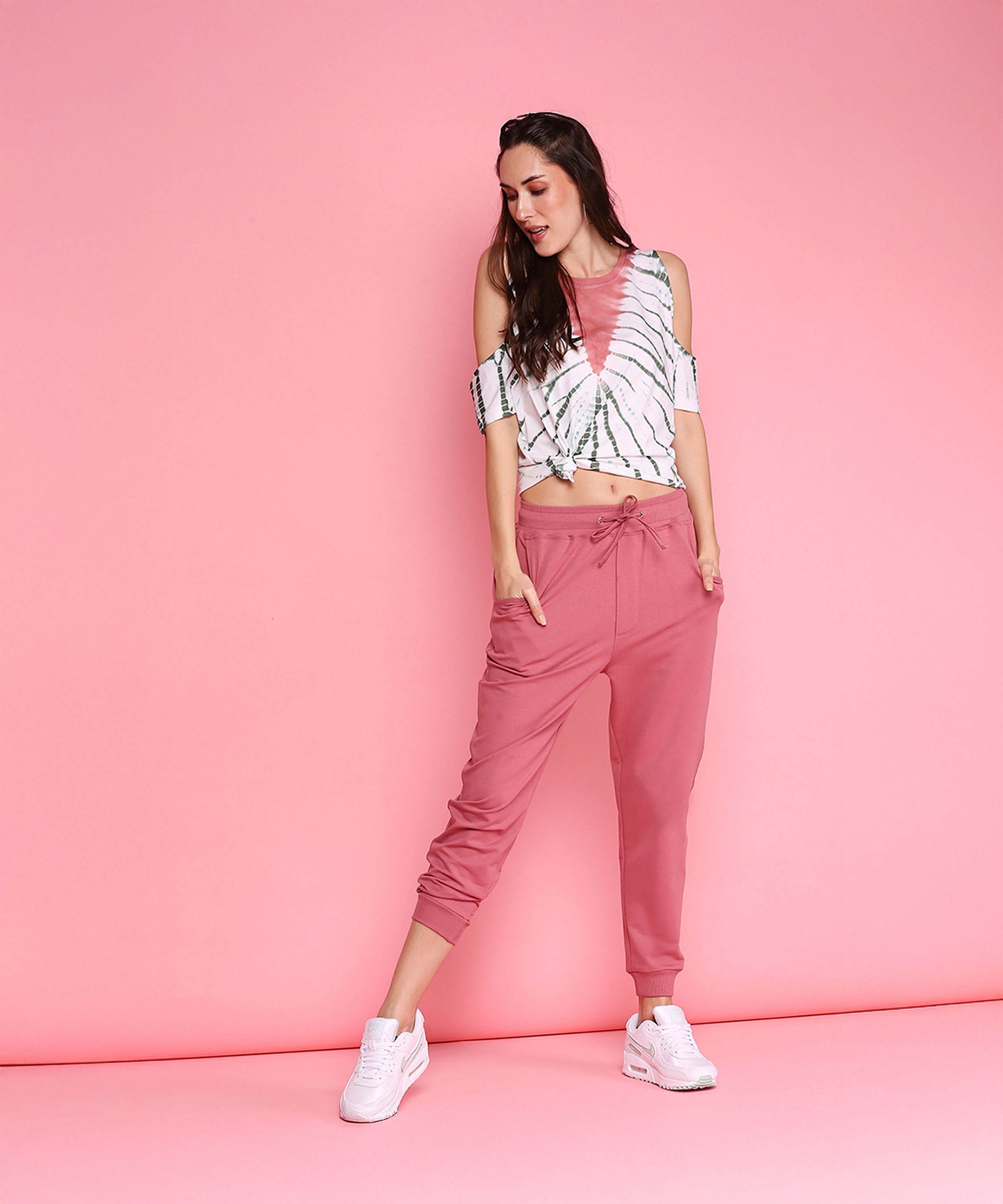 Woman models the Cold Shoulder Comfort Fit Top + Joggers - AW21 in carrot pink, paired with white sneakers, posing confidently against a pink background.