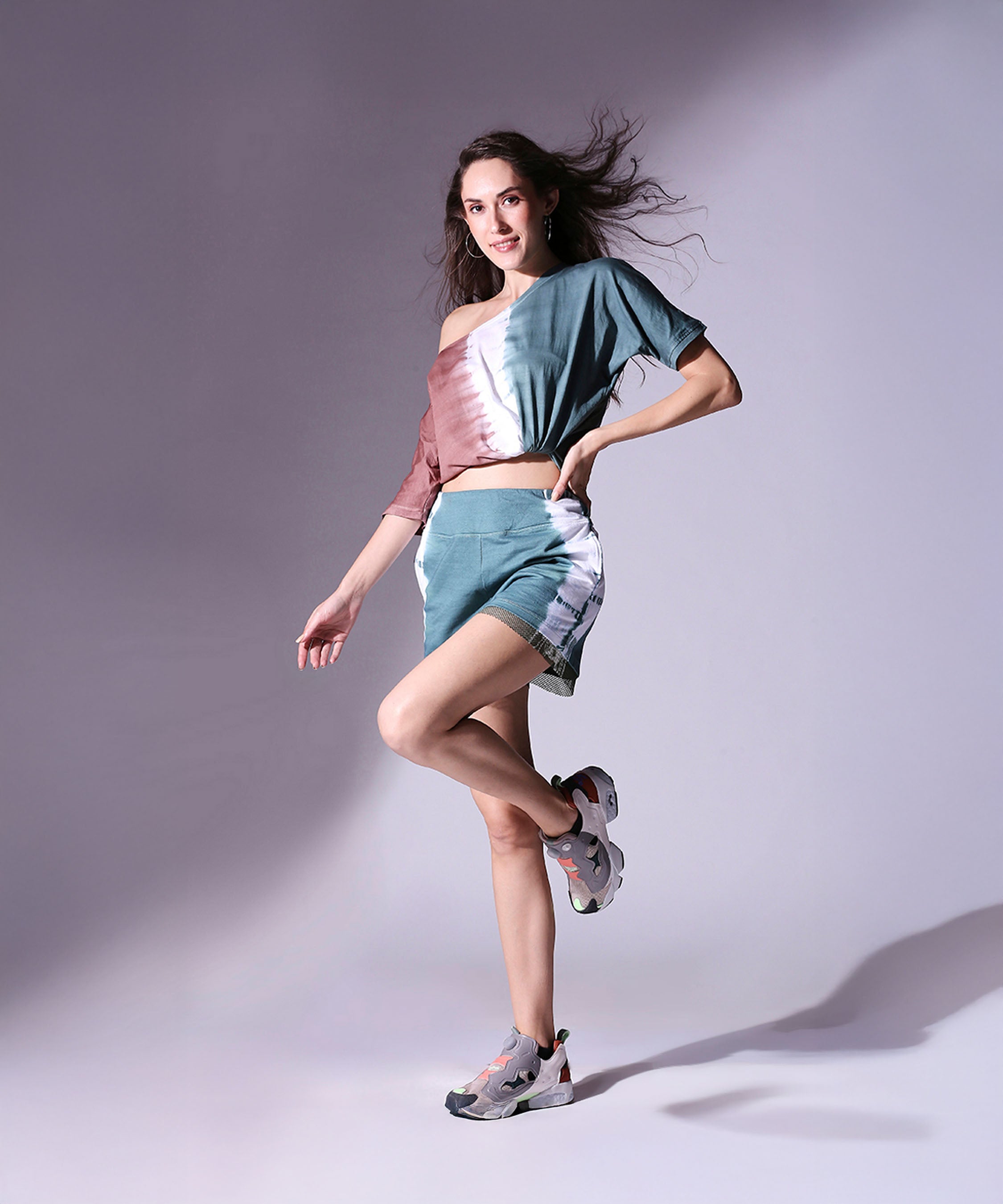 Woman models the Oversized Comfort Tee + Shorts - AW21 set, paired with sneakers, against a softly lit plain background.