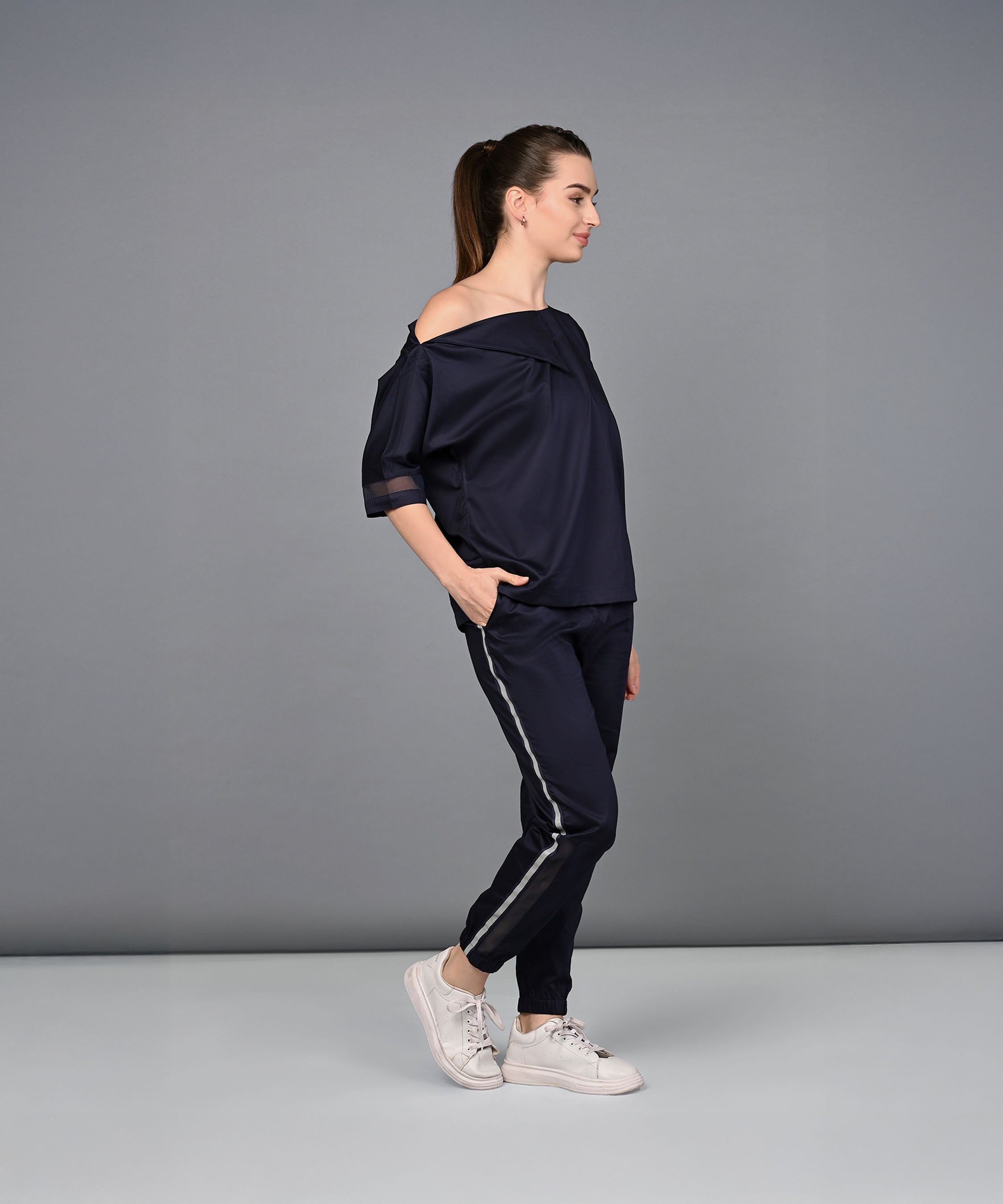 A woman stands sideways with hands in pockets, wearing the Off Shoulder Nebula Navy Co-ord Set made from 100% cotton, featuring side-striped pants and a relaxed fit.