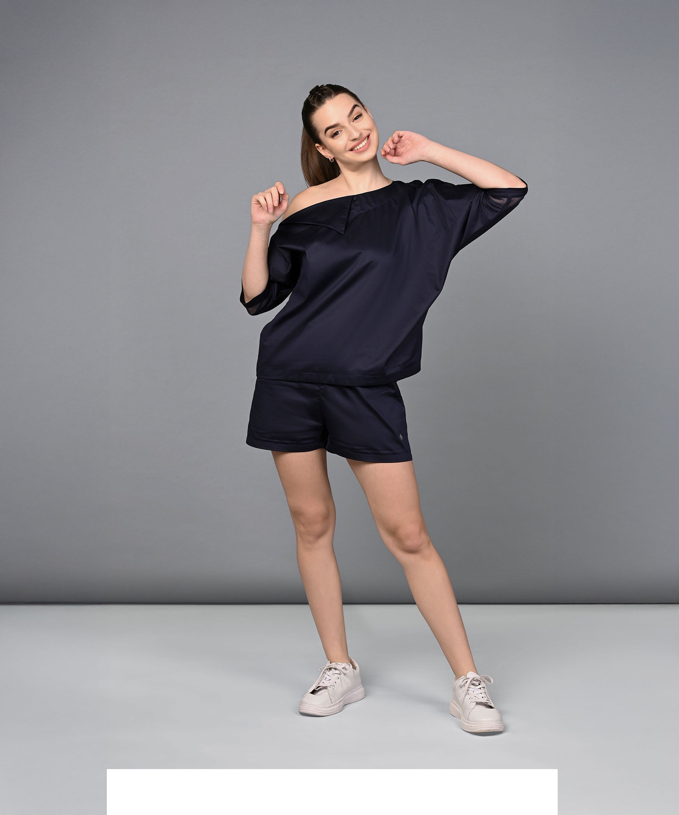 A woman cheerfully poses against a plain gray studio background in the Off Shoulder Nebula Navy Co-ord Set, crafted from 100% cotton.