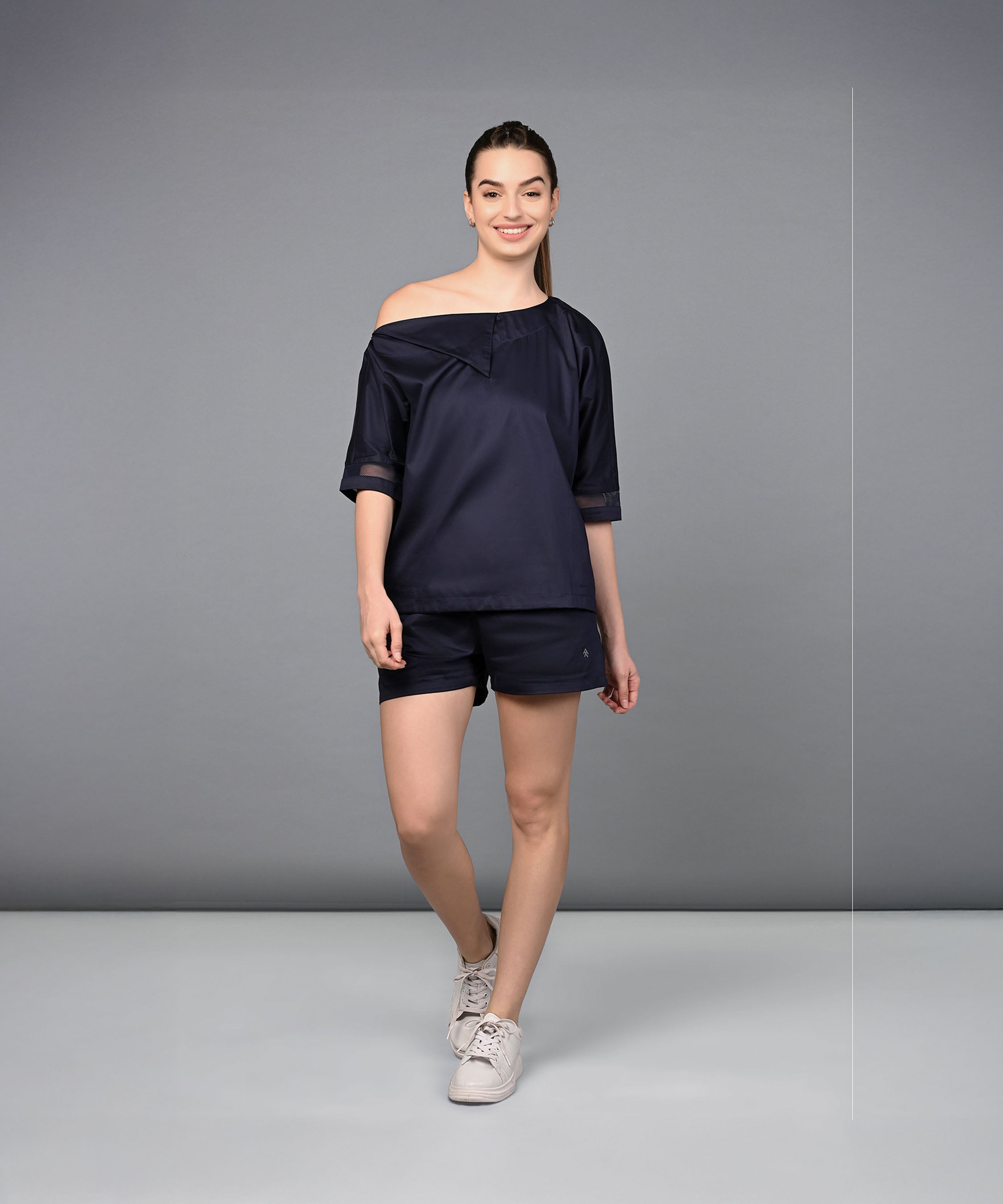 A woman smiles wearing the Off Shoulder Nebula Navy Co-ord set, crafted from 100% cotton, as she poses against a plain gray background.