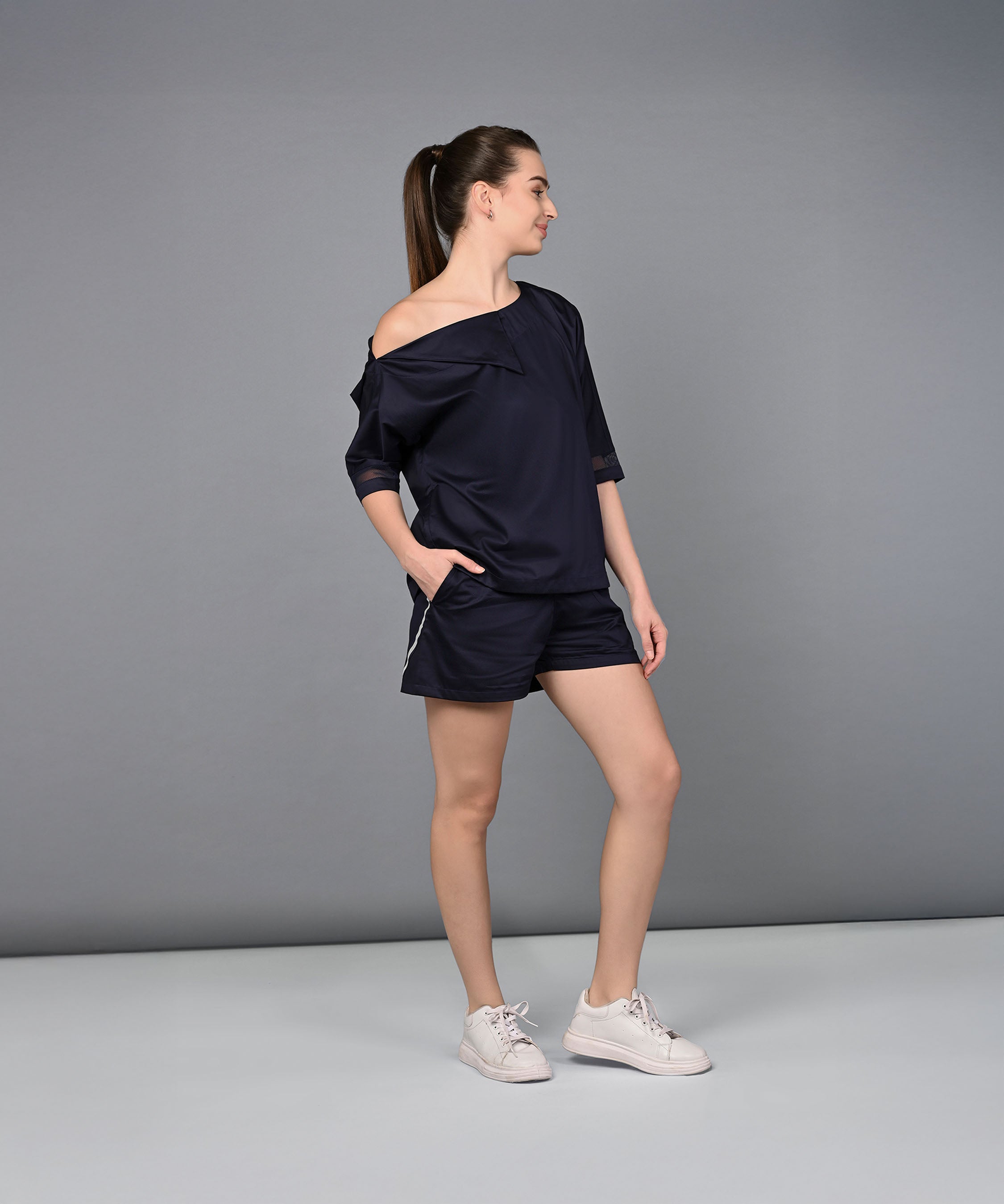 A woman stands sideways, hands in pockets, wearing white sneakers and the Off Shoulder Nebula Navy Co-ord set crafted from 100% cotton.