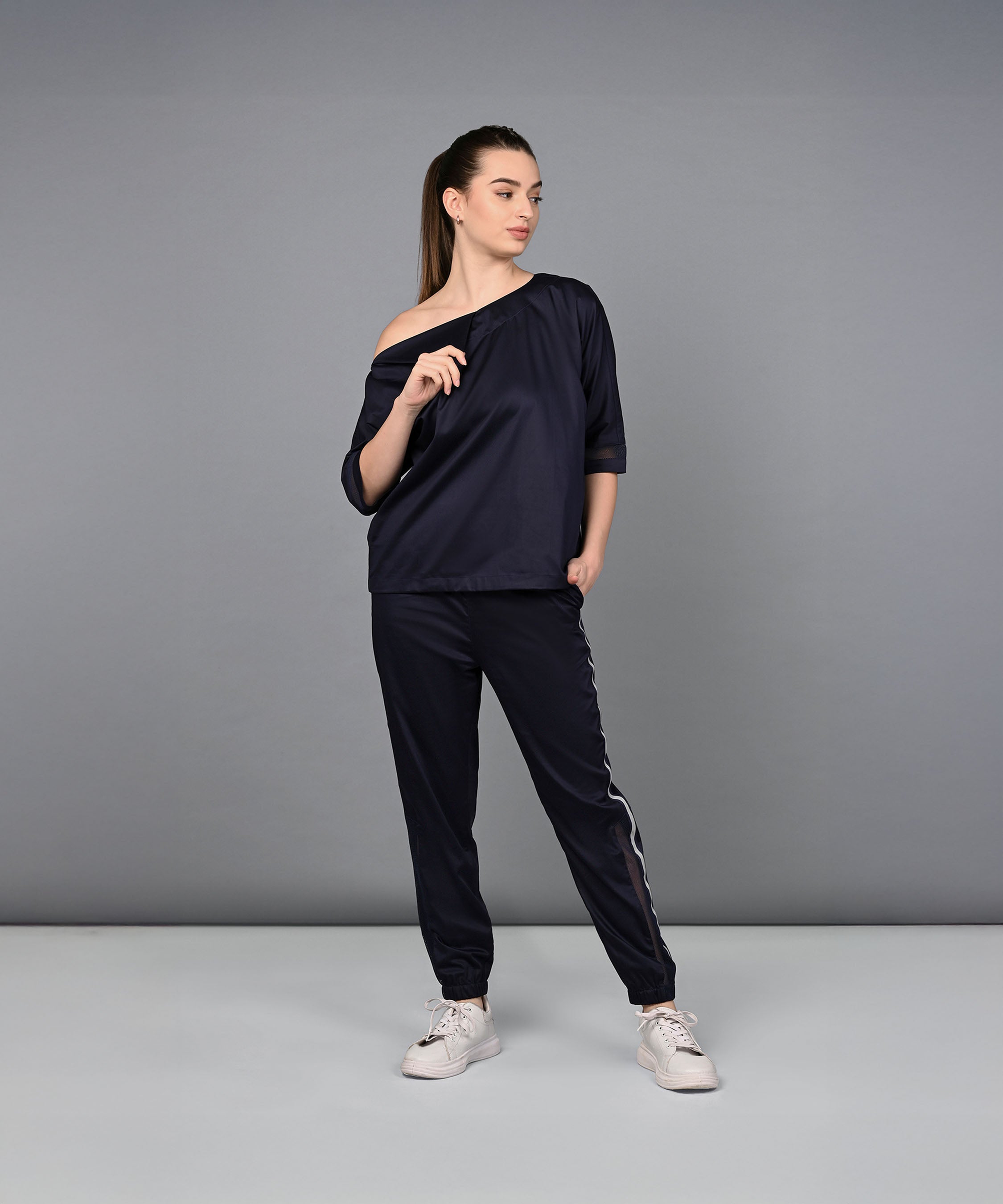 A woman in the Off Shoulder Nebula Navy Co-ord Set, crafted from 100% cotton, stands with one hand in her pocket and points sideways while wearing white sneakers.
