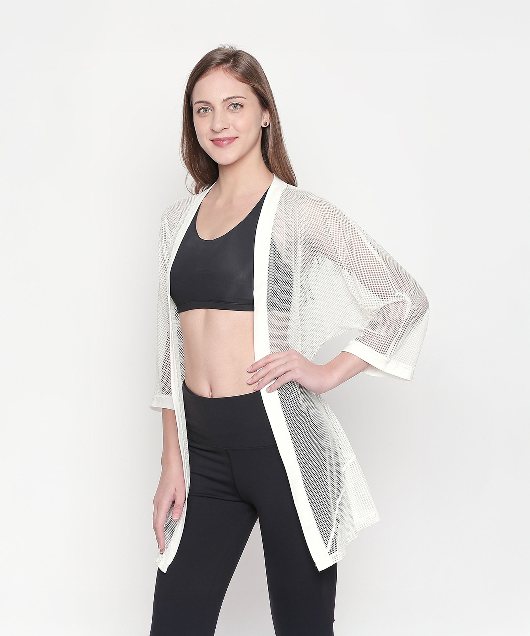 Woman models the Loose Fit 3/4th Sleeve White Shrug for Women over a black sports bra and leggings, posing and smiling to showcase an effortless athleisure style.