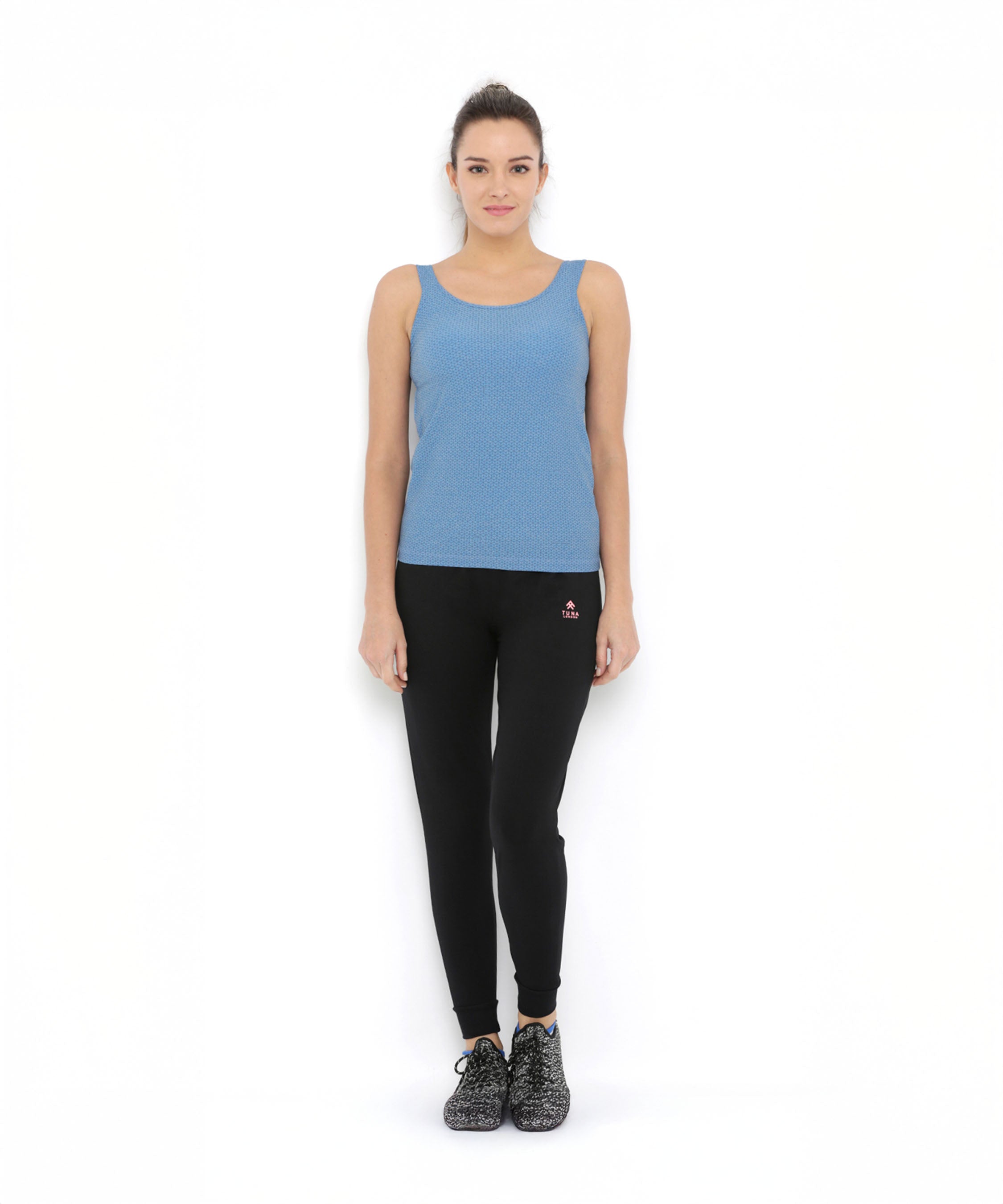 A woman stands against a white background wearing a blue tank top and Black Classic Joggers TLC02, featuring a slim tapered fit, paired with black shoes.