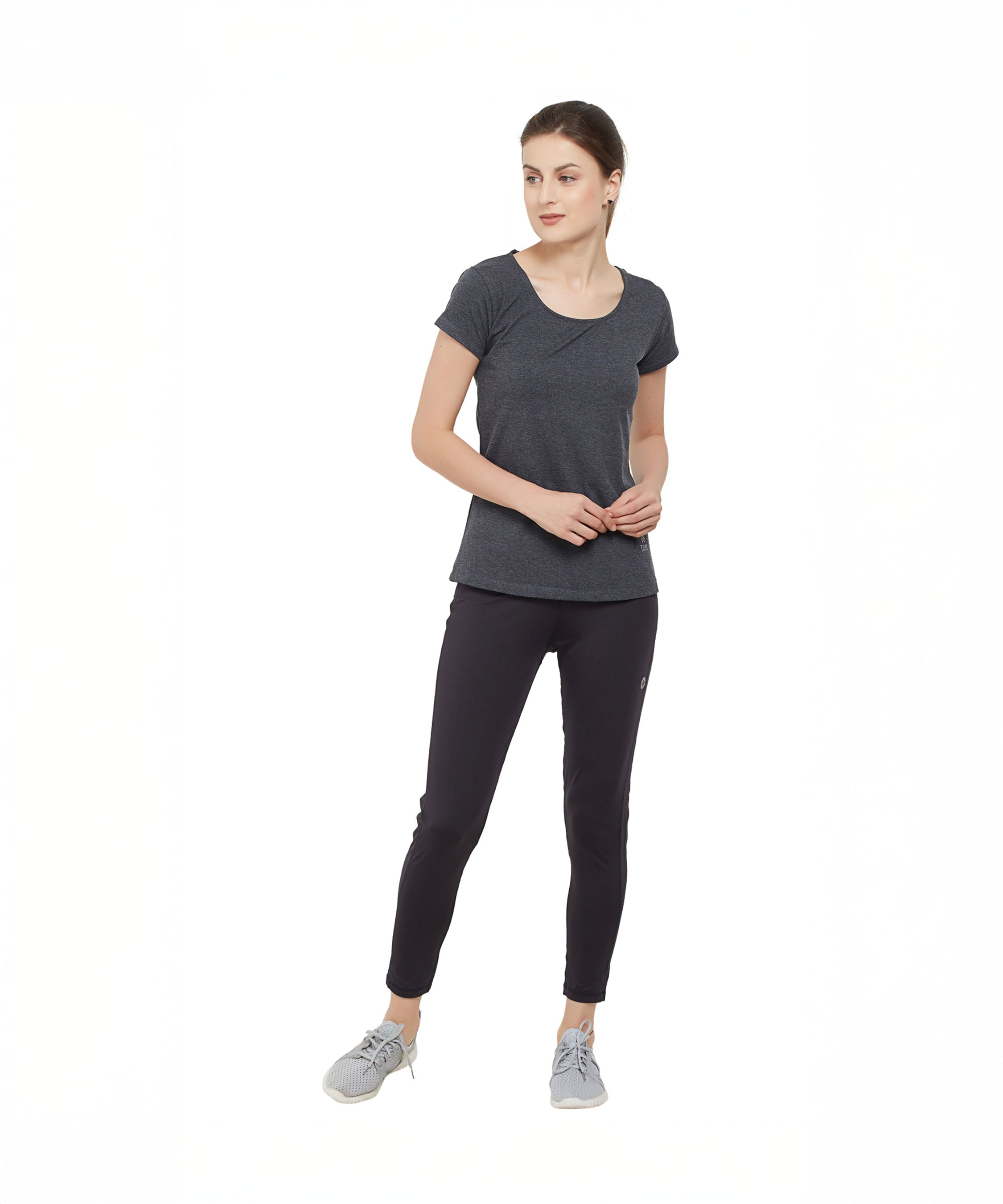 Woman in a TUNA LONDON Grey Round Neck Tee, black leggings, and gray sneakers standing against a white background.