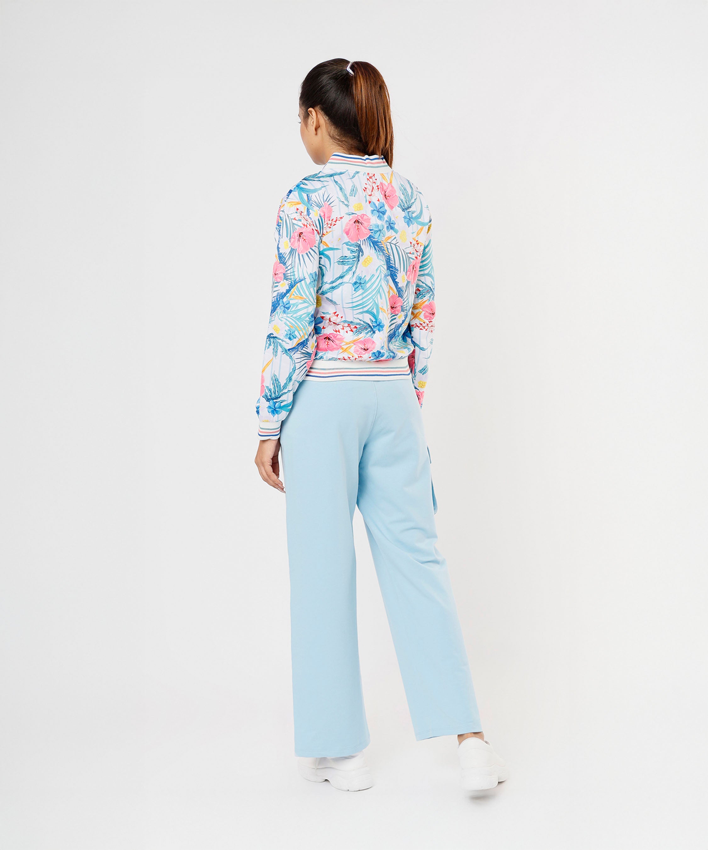 A woman faces away wearing the Blossom Blue Set with Crop Top—floral bomber jacket, light blue cargo track pants, and white shoes—against a white background.