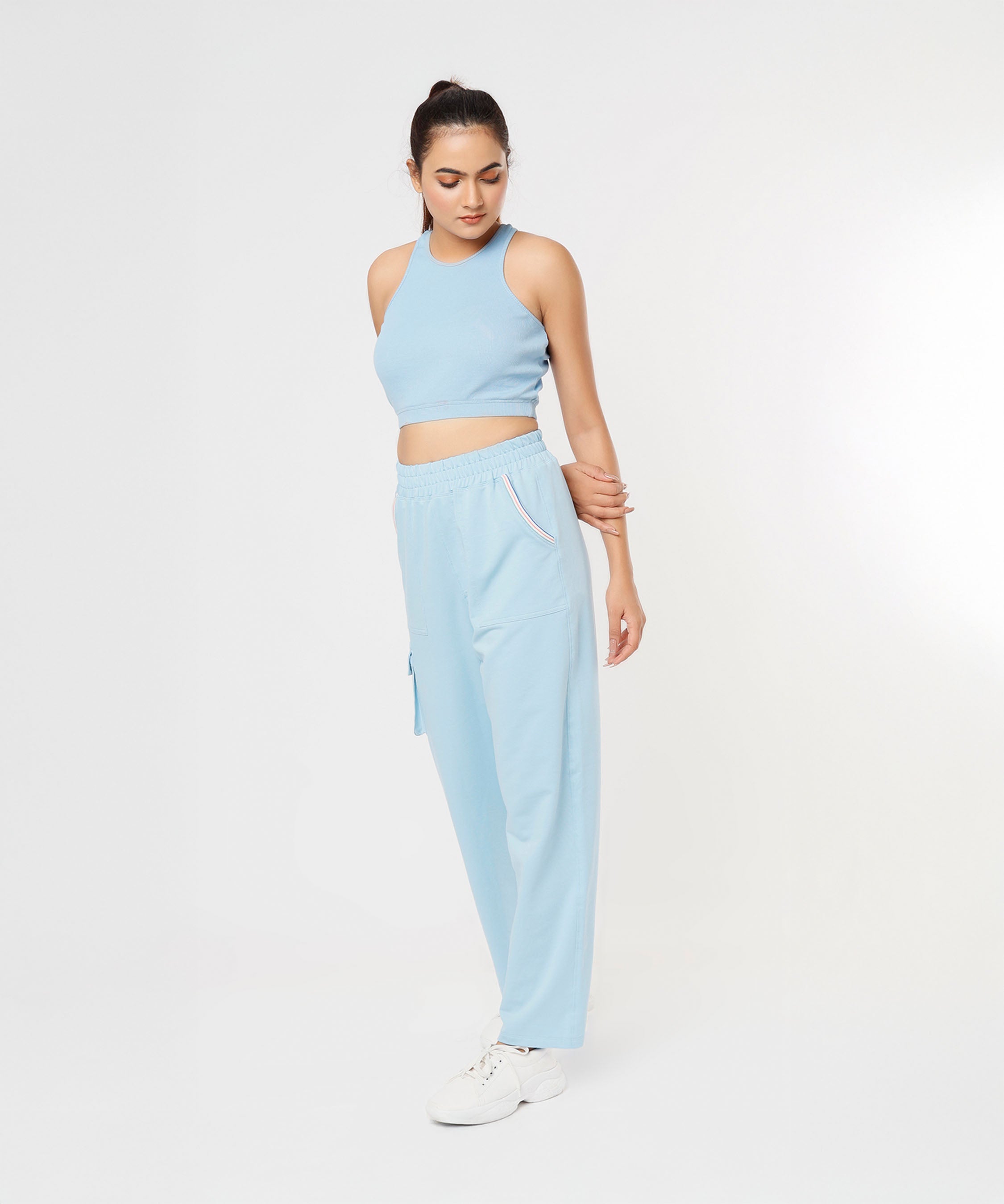 Woman wearing the Blossom Blue Set with a light blue sleeveless crop top and cargo track pants, standing and looking down against a white background.