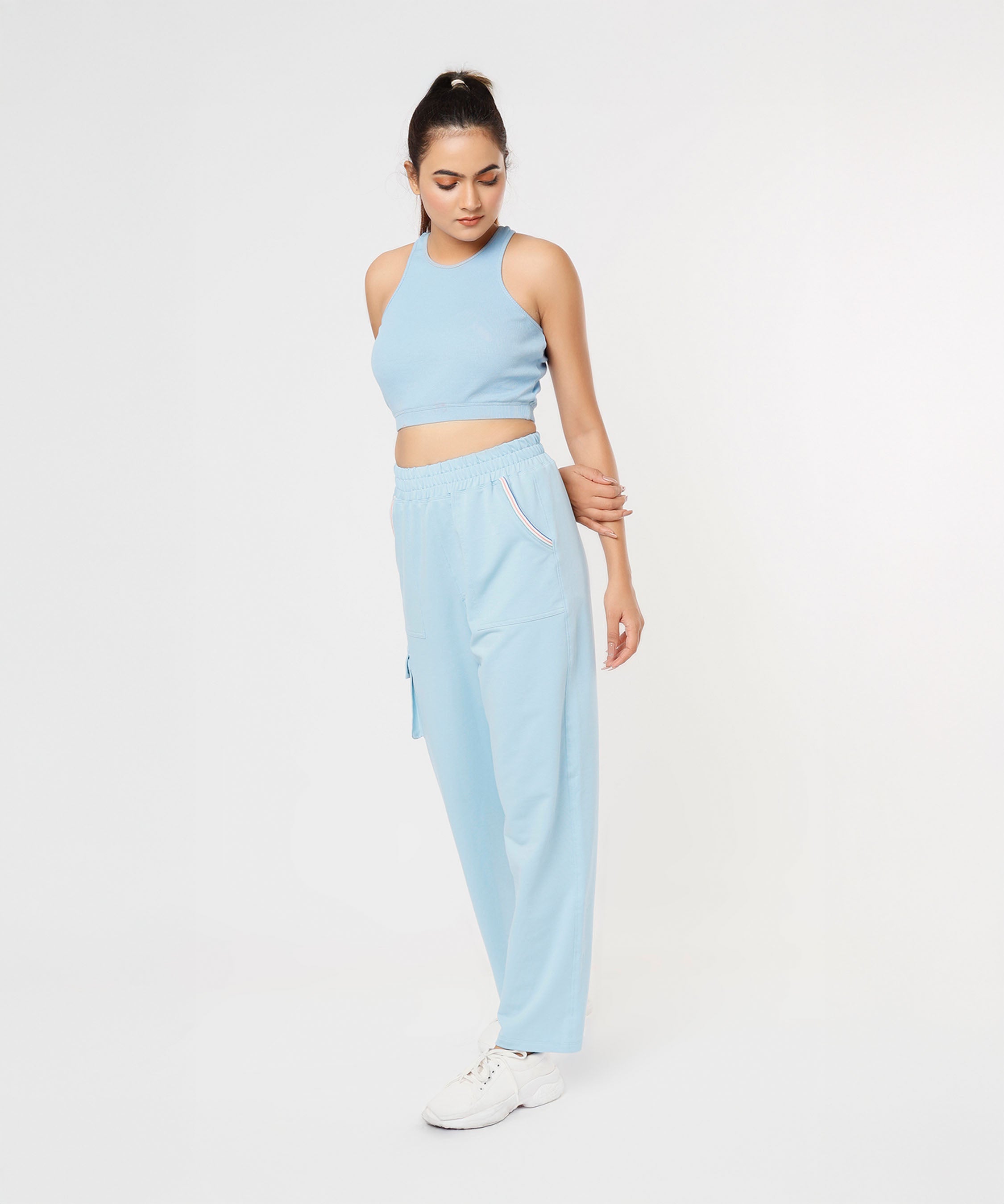 Woman wears the Blue Dream Crop Top—a powder blue racerback athleisure piece—with matching joggers and white sneakers, standing while looking down.