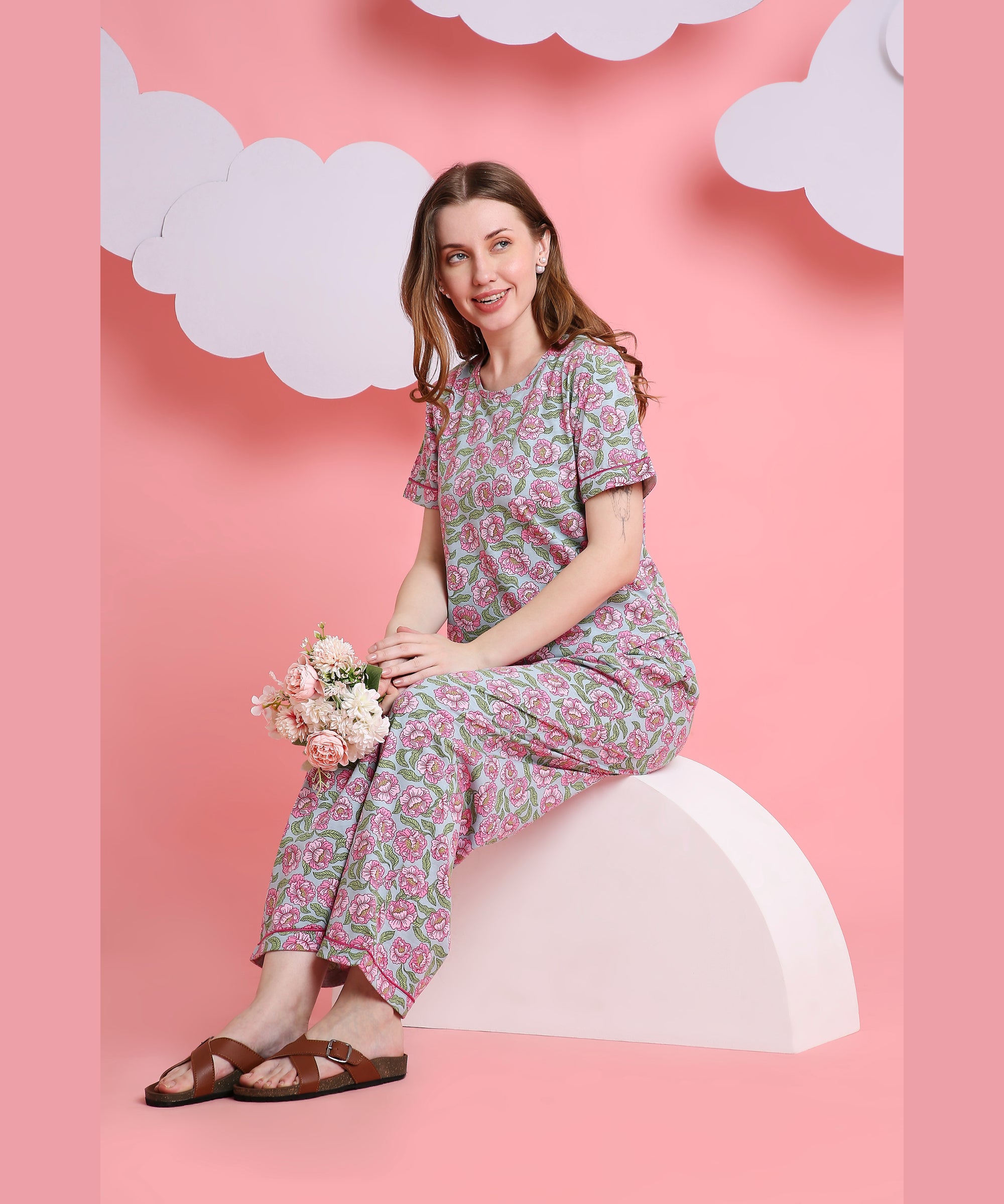 A woman in the Floral Love pink floral night suit sits on a white arch, holding flowers amid clouds on a pink background—showcasing this comfortable sleepwear made from breathable fabric.