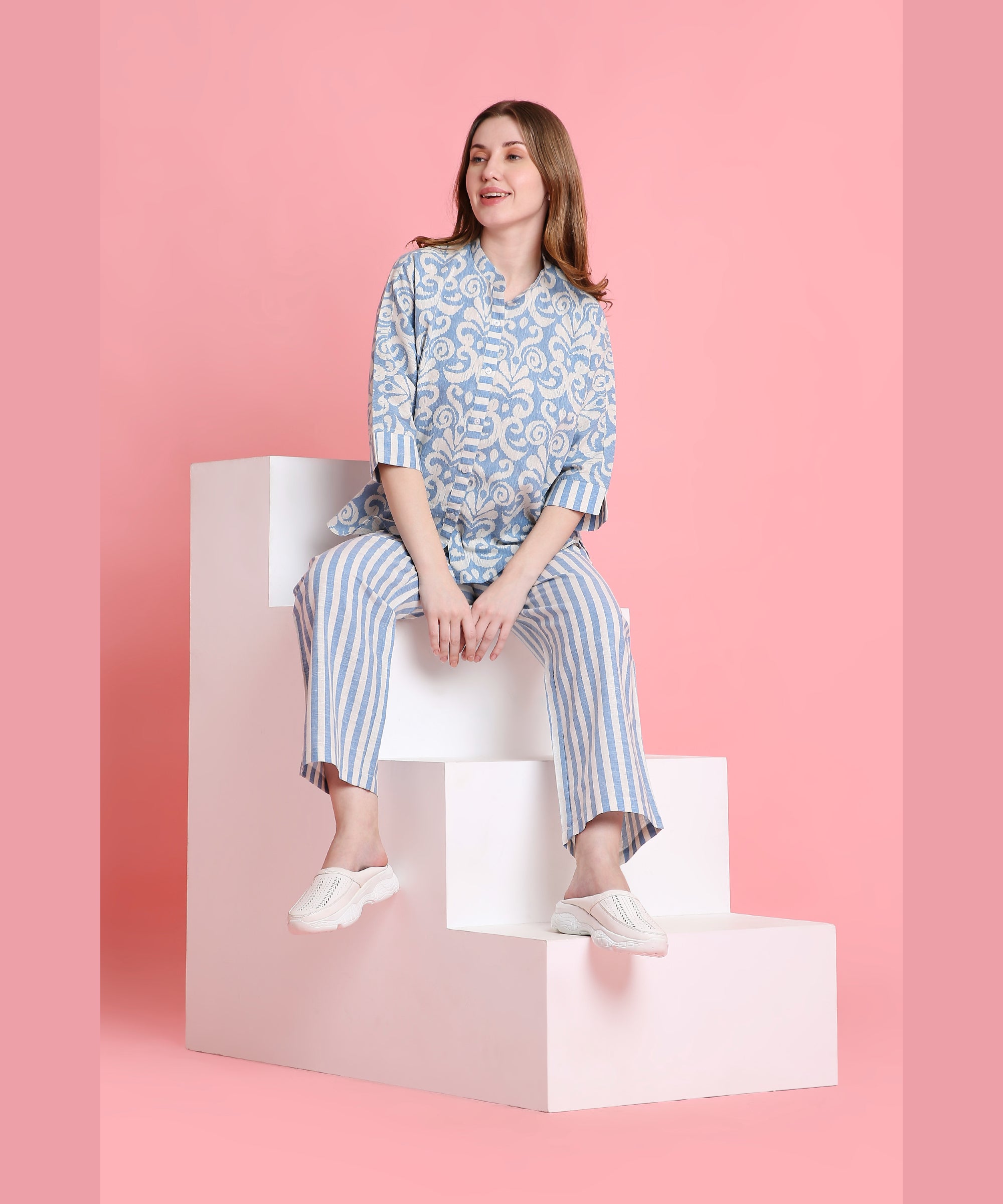 A woman wearing the Linen Blue pyjama set sits on white blocks against a pink background, smiling.