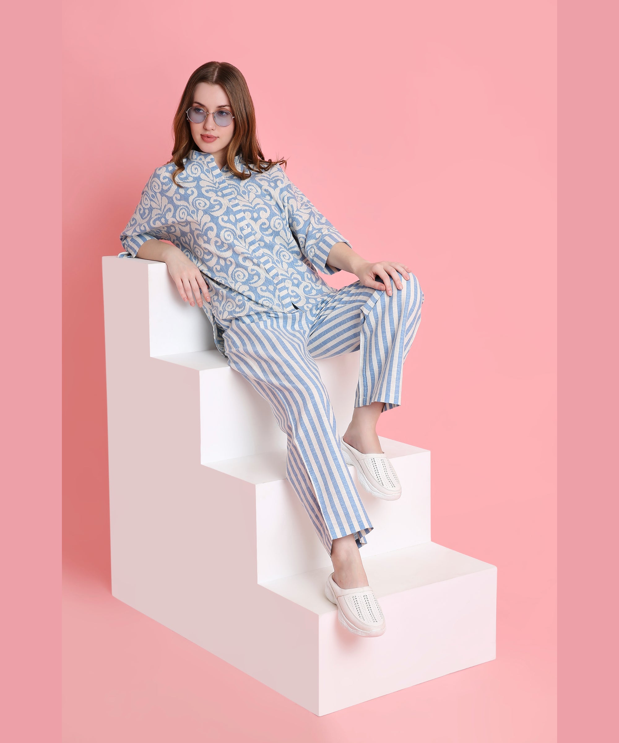 A woman in Linen Blue damask-print top and relaxed-fit pyjamas sits on white steps with a pink background, wearing sunglasses.