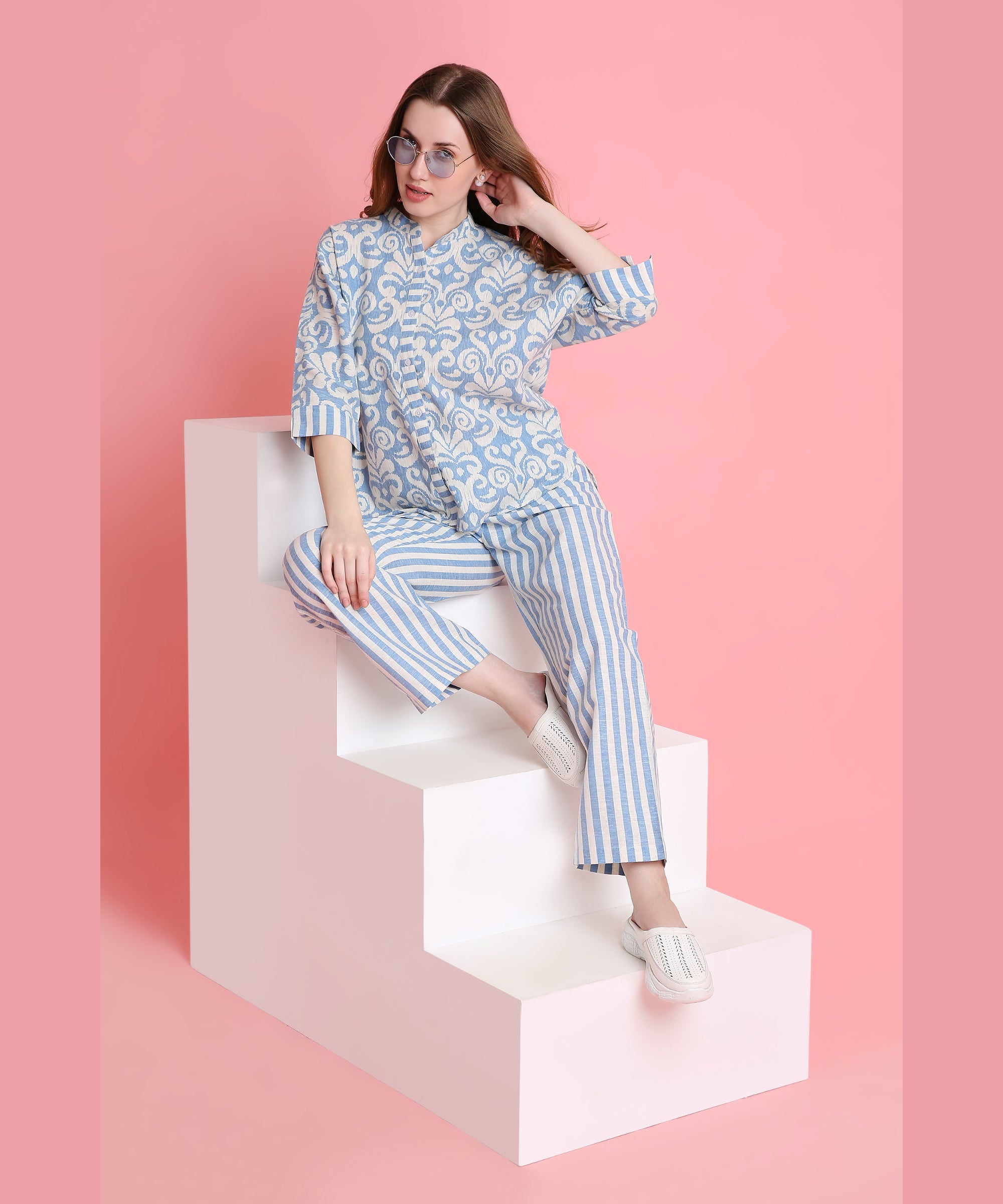 A woman in the Linen Blue damask-print top and relaxed-fit pyjamas sits on white steps against a pink background, wearing sunglasses.