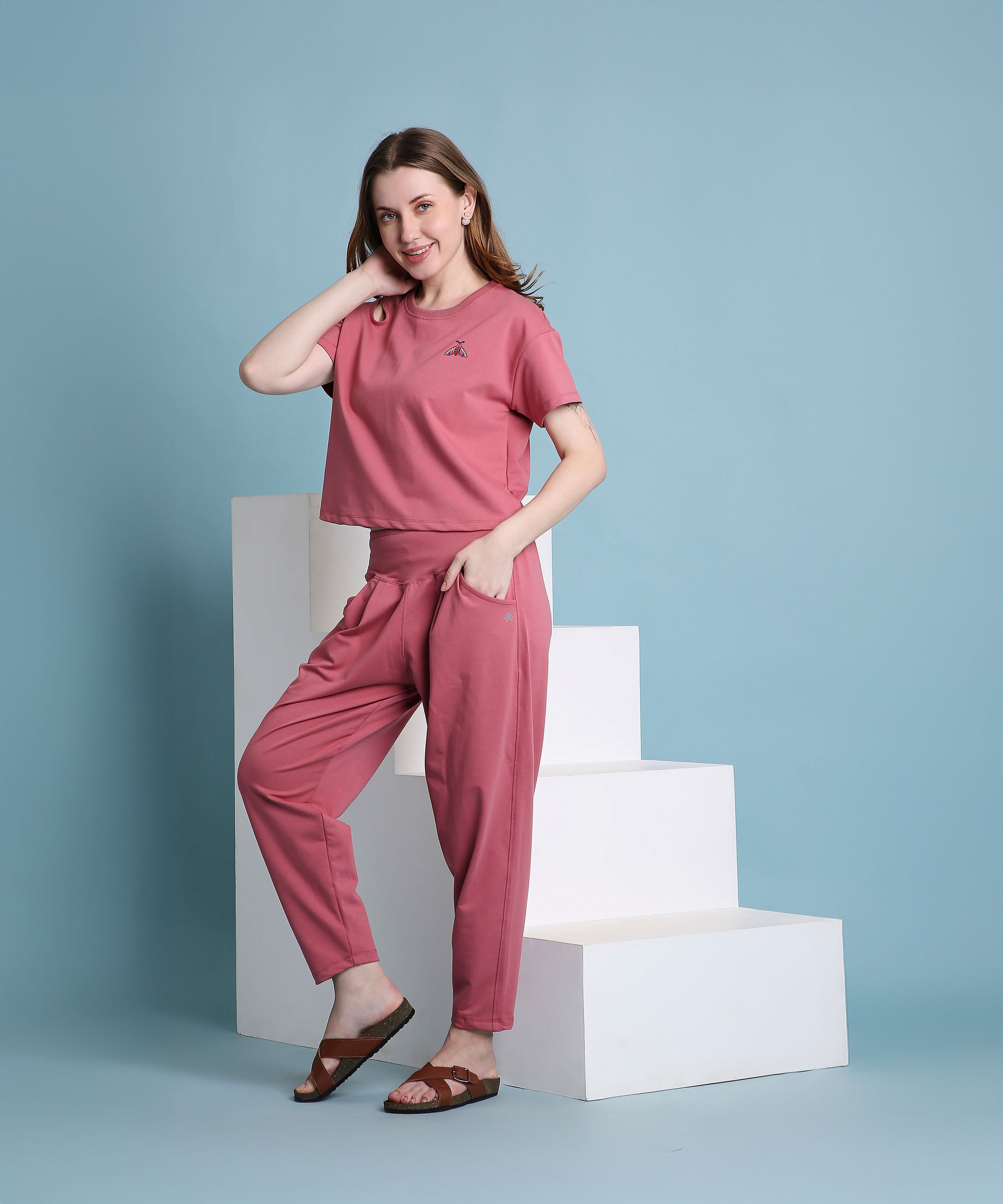 A woman wearing the Vintage Pink Knit Cord Set poses by white steps, smiling with one hand on her hip and the other near her face.