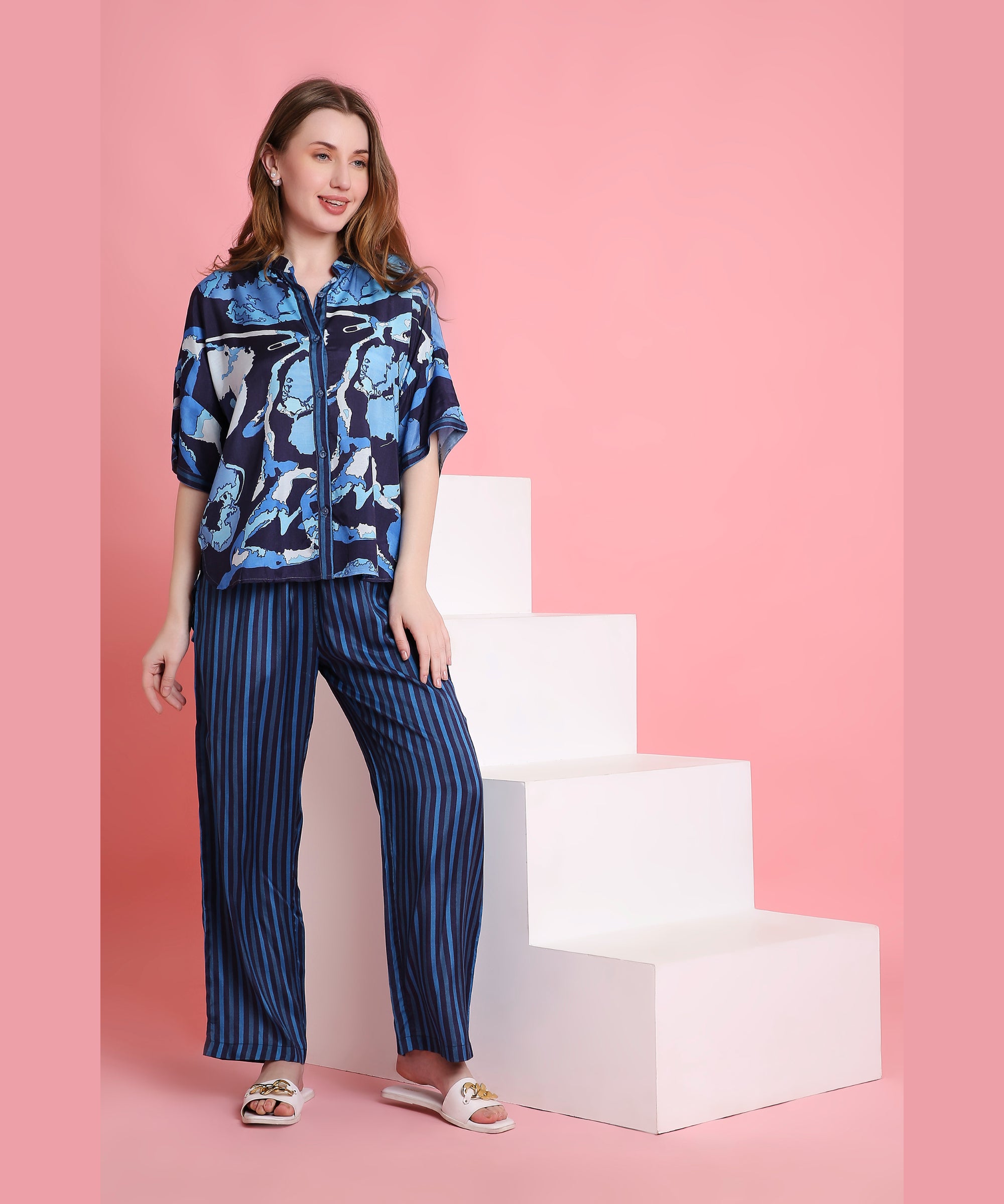 A woman in Ocean Breeze loungewear—a blue patterned shirt and striped pants—stands by white steps on a pink background, highlighting the set’s comfy, breathable fabric.