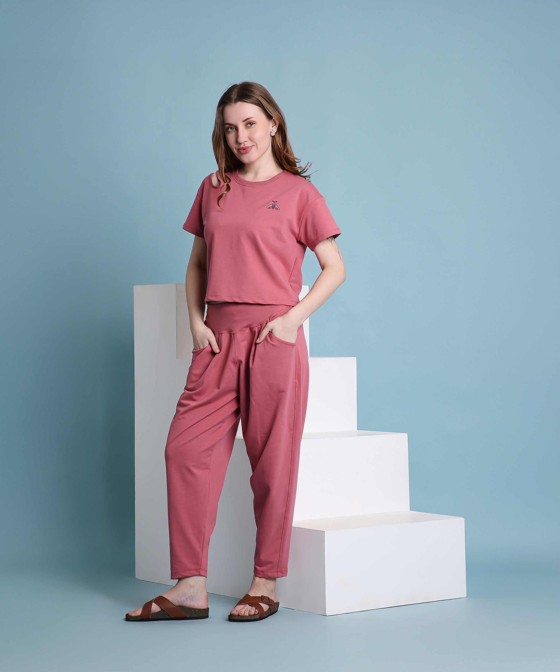 A woman, standing by white steps against a blue background, wears the Vintage Pink Knit Cord Set with pleated yoga pants.