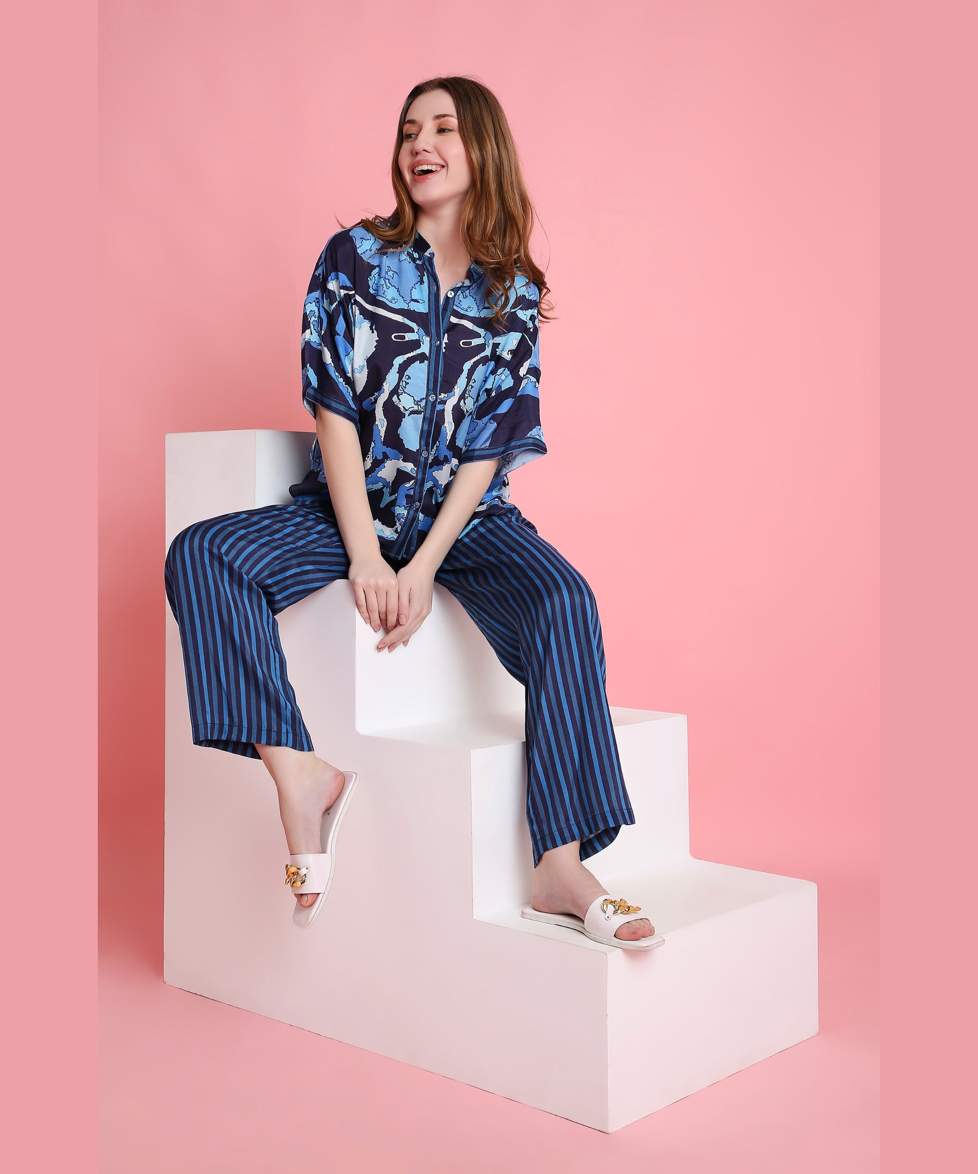 Smiling woman in the Ocean Breeze blue patterned shirt and striped pants, made from breathable fabric, sits on white steps with a pink background.