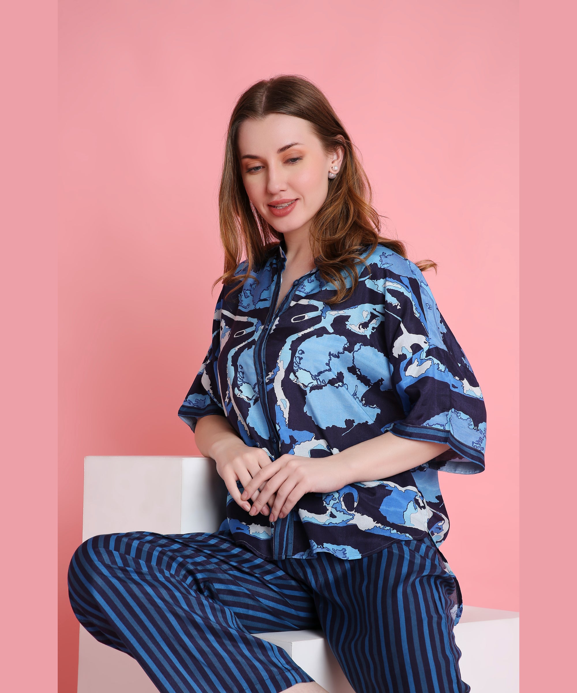 Smiling woman in an Ocean Breeze blue patterned top and striped pants sits against a pink background—showcasing comfy, breathable loungewear.