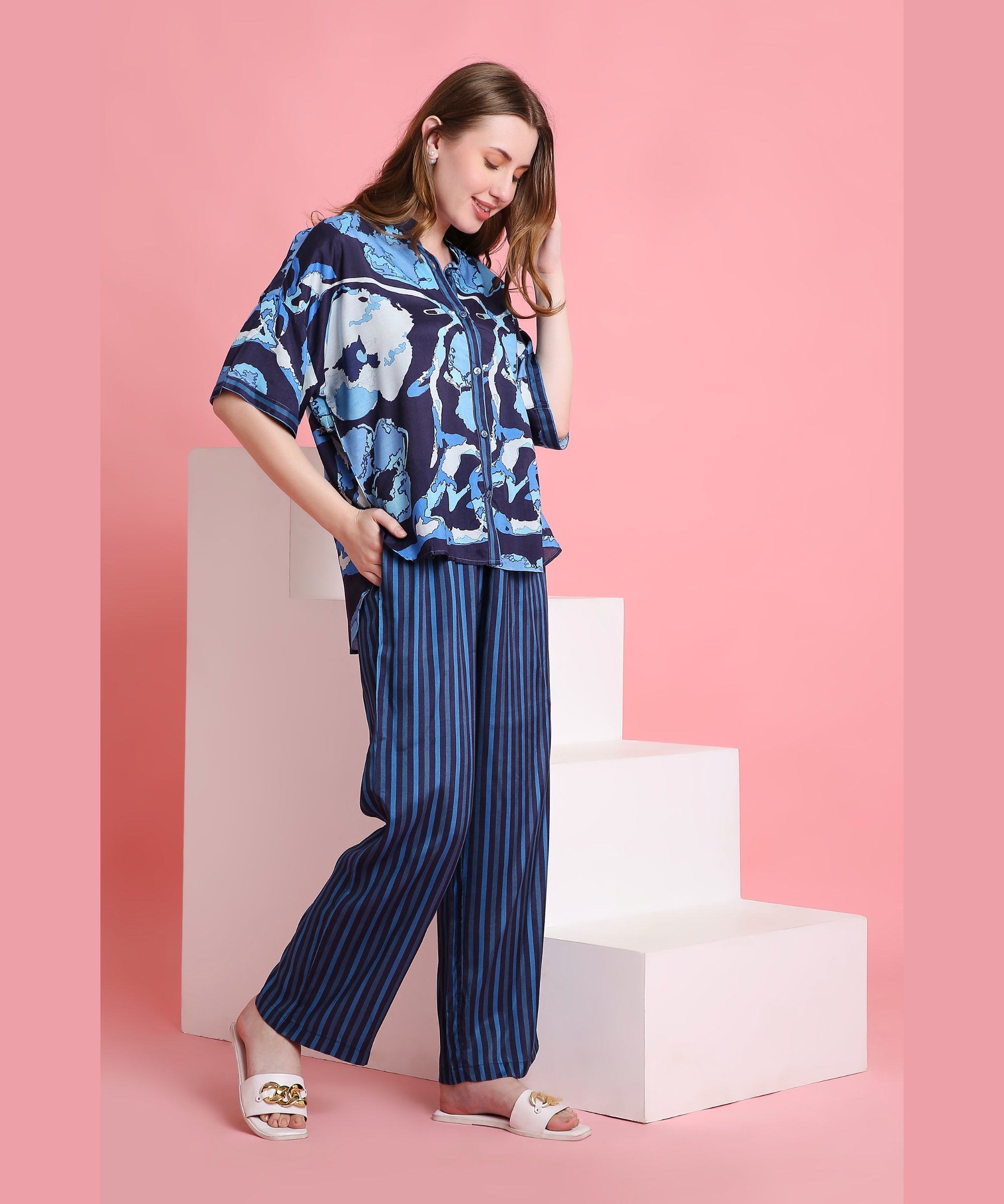 Wearing the Ocean Breeze blue floral top and striped pants made from breathable fabric, a woman stands by white steps with a pink background, smiling downward.