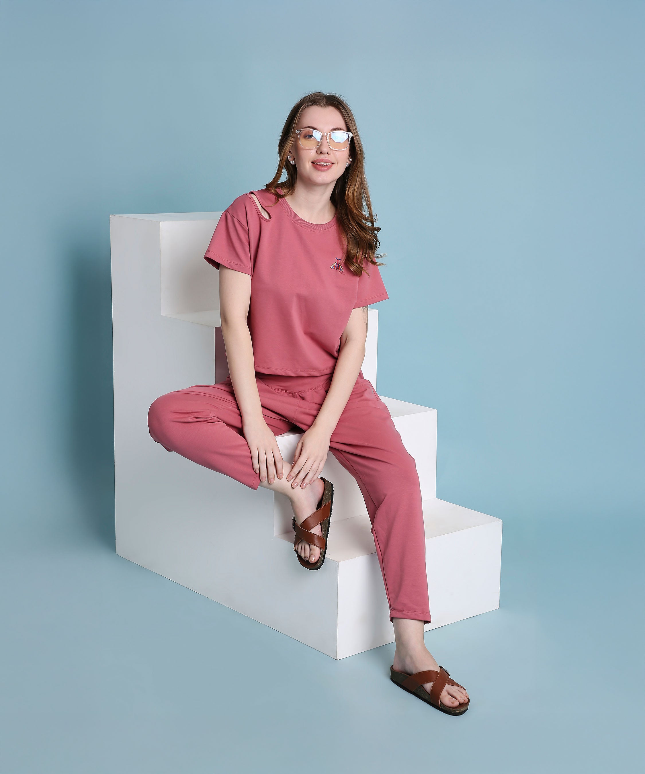 A woman wearing a Vintage Pink Knit Cord Set and sandals sits on white steps against a blue background, smiling at the camera.