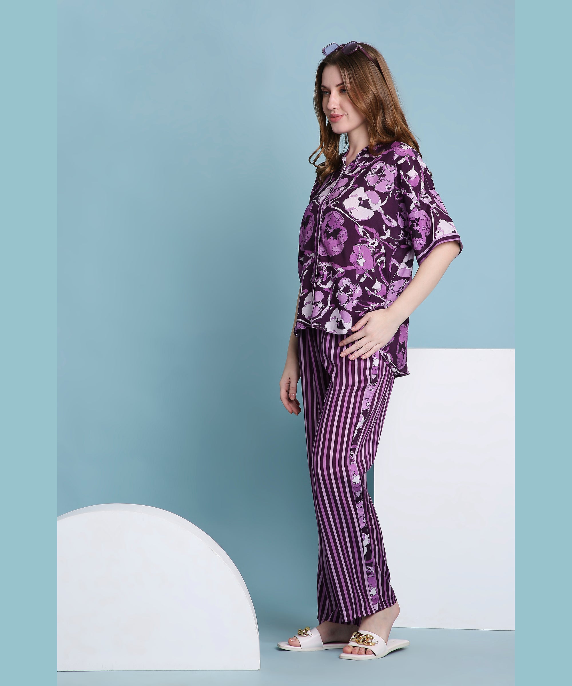 A woman models the Purple Breeze pyjama set—featuring floral and striped designs—while standing sideways against a blue background in white sandals, showcasing a fresh, stylish approach to women’s loungewear.