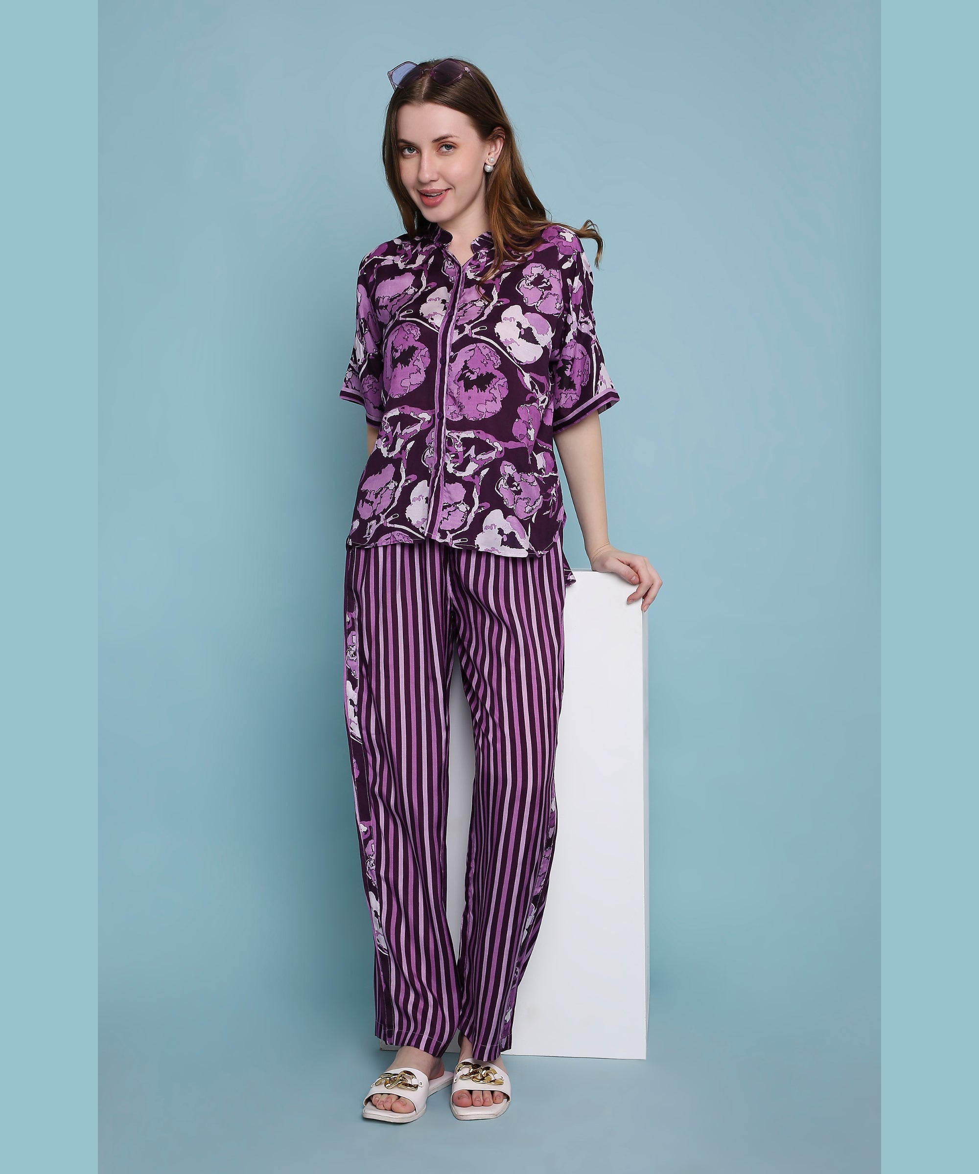 A woman smiles in comfortable loungewear, wearing the Purple Breeze pyjama set with matching purple floral and striped patterns, standing against a blue background.