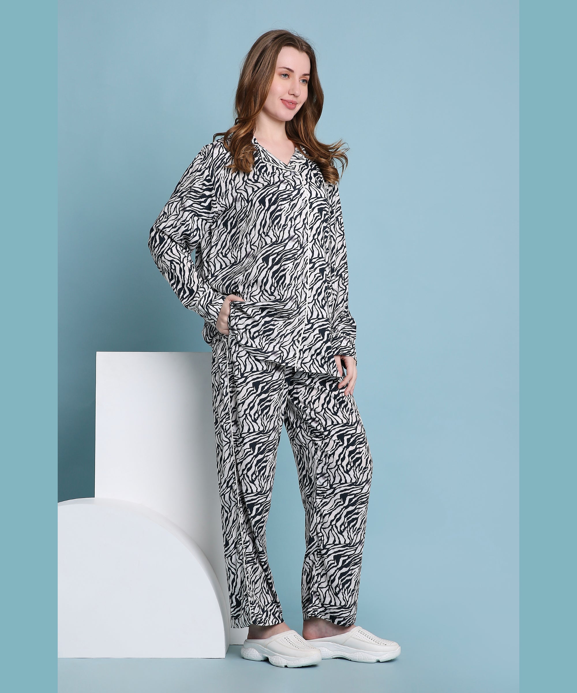 A woman stands against a blue backdrop with geometric props, wearing the relaxed fit Zeba zebra print pyjama set and white shoes.