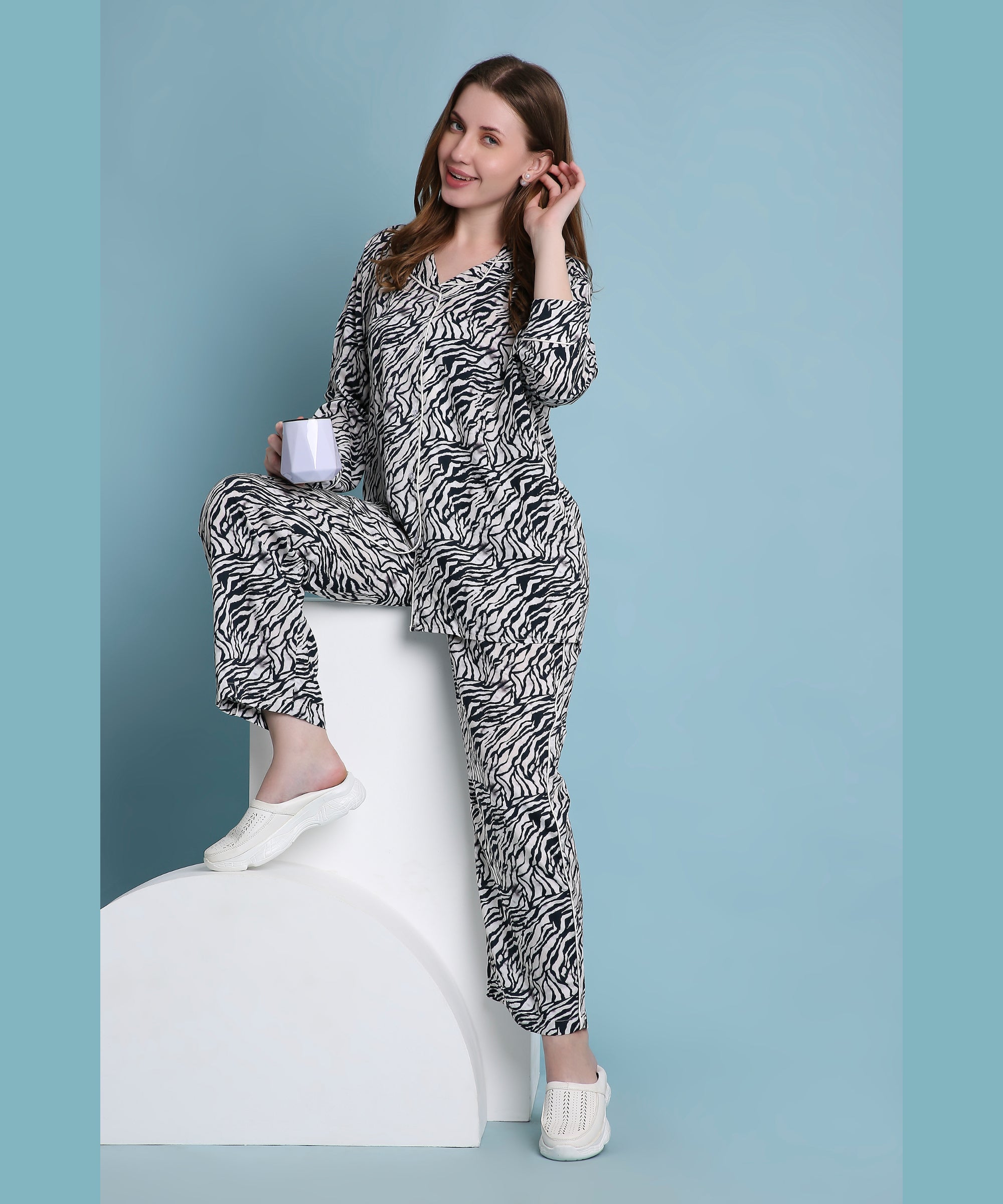 A woman in the Zeba relaxed fit zebra print pyjama set stands on a white block, smiling with a mug against a blue background—perfect for stylish loungewear.