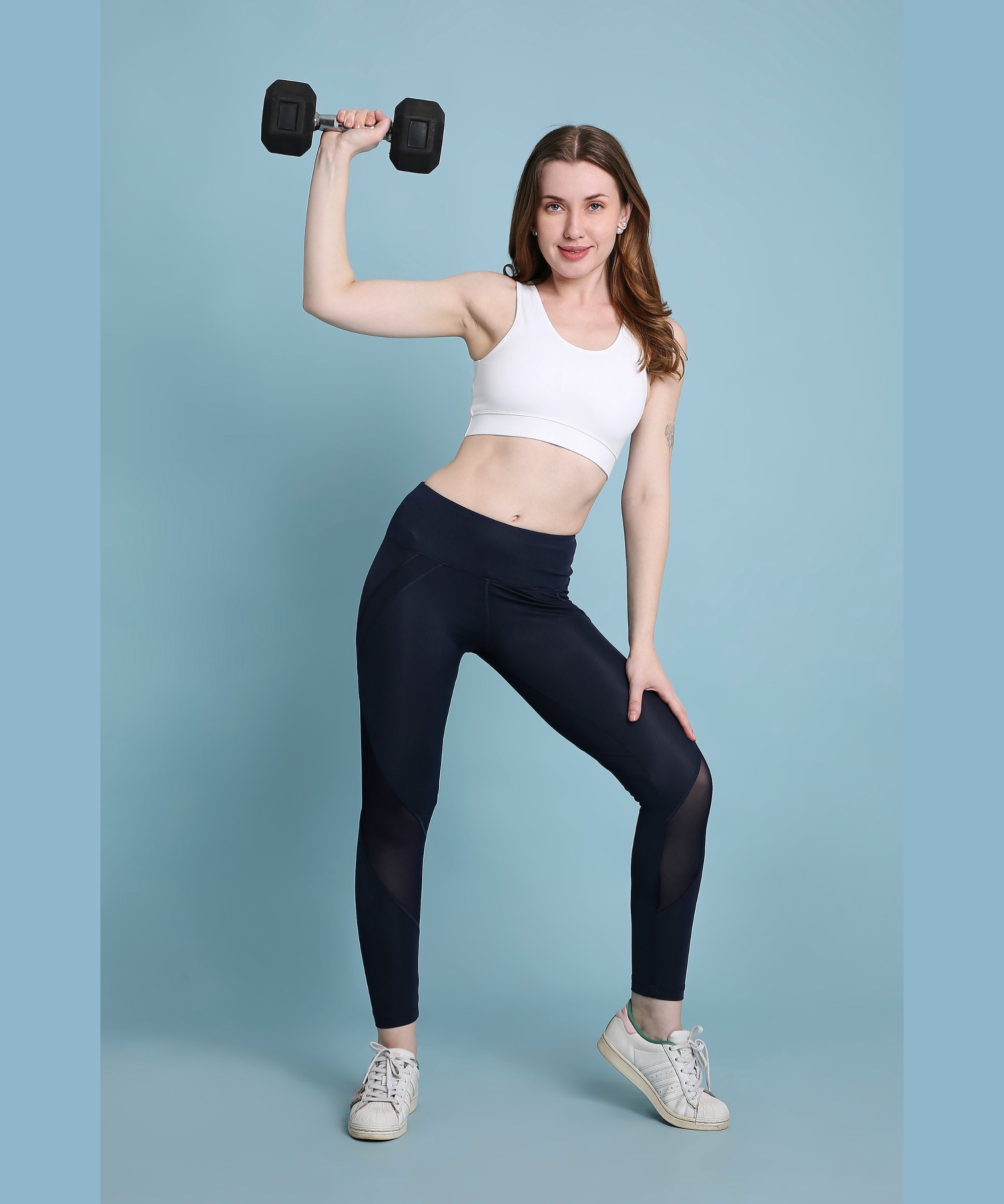 A woman in athletic wear smiles while raising a dumbbell against a blue background, highlighting the stylish Indigo poly blended leggings.