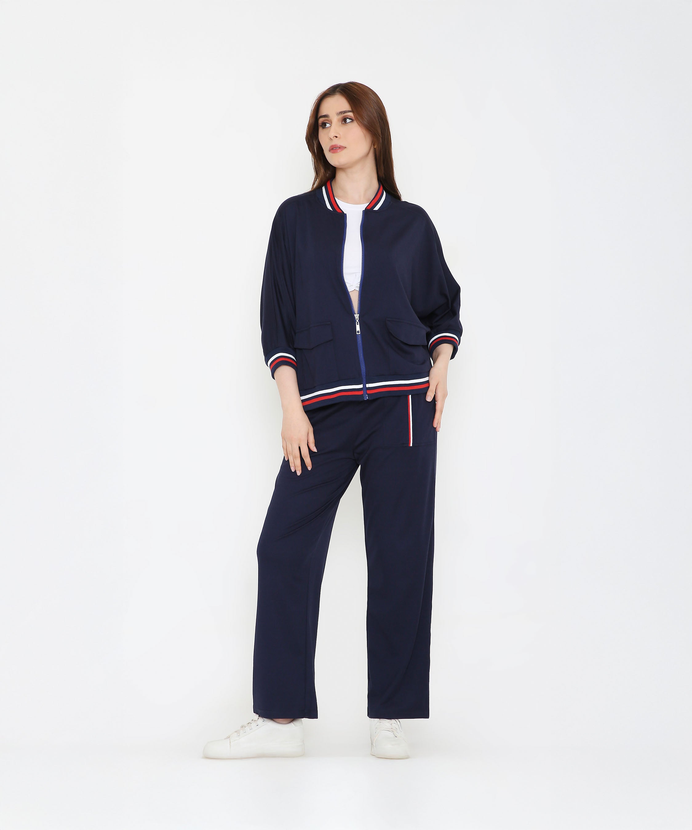 A woman in a Navy Blue Zipped Long Jacket with Tracks and white sneakers stands with one hand in her pocket against a white background.