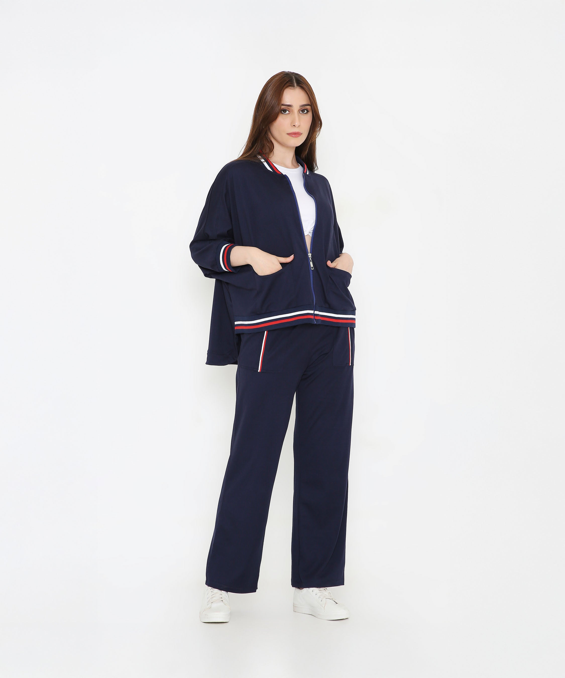 Woman in a Navy Blue Zipped Long Jacket with matching tracks, accented by red and white stripes. Features pockets for convenience—stylish women's activewear ideal for comfort and paired perfectly with white sneakers.