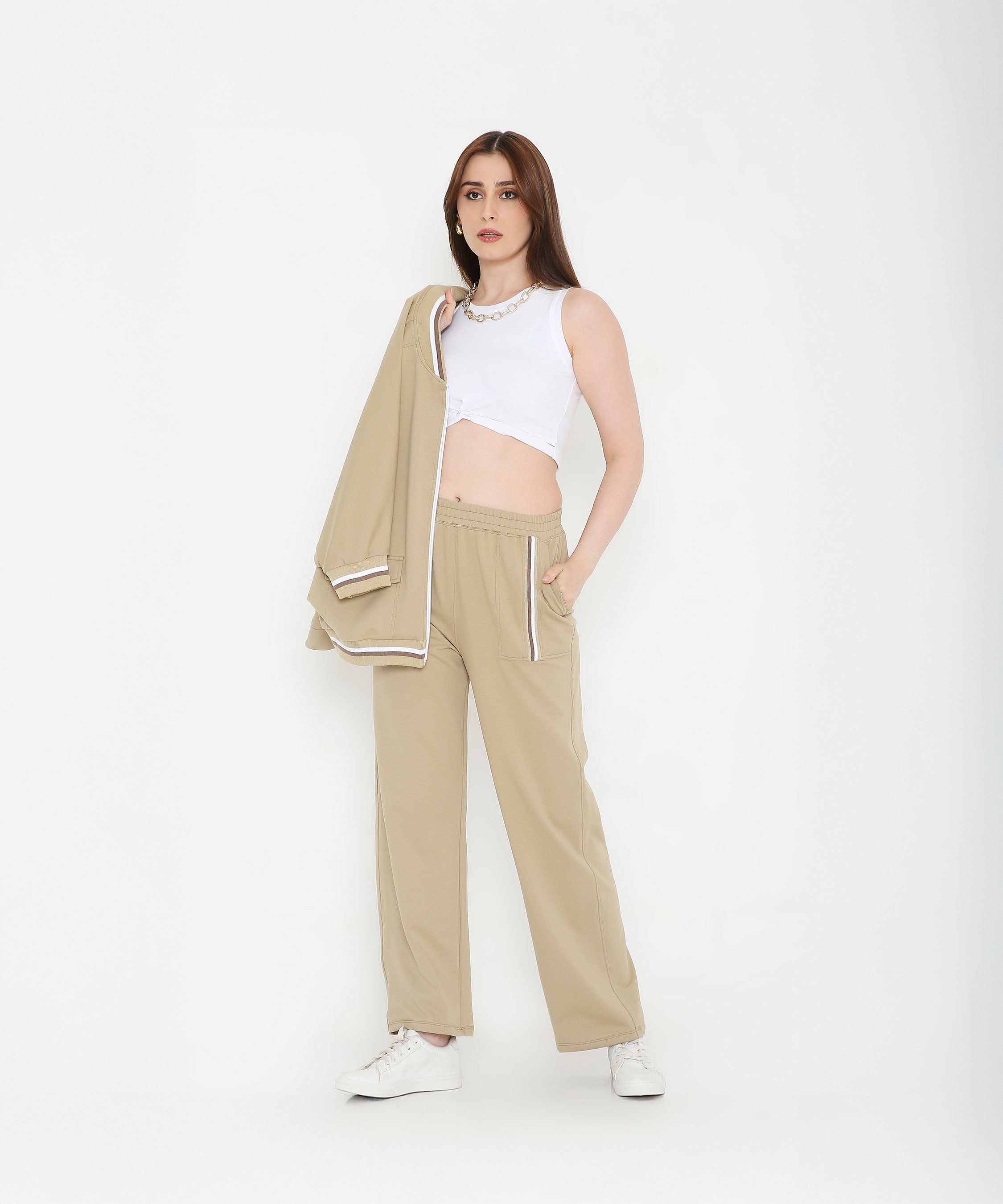 A woman in beige pants and white crop top holds the Beige Zipped Long Jacket with Tracks, standing against a white background—a modern, versatile track set for women.