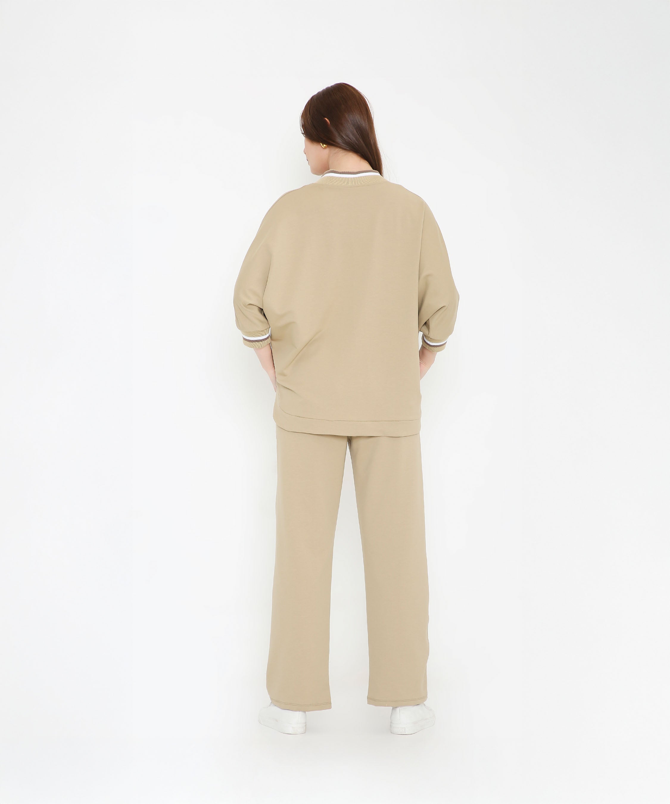 Woman facing away, dressed in the Beige Zipped Long Jacket with Tracks and white sneakers, standing against a white background.