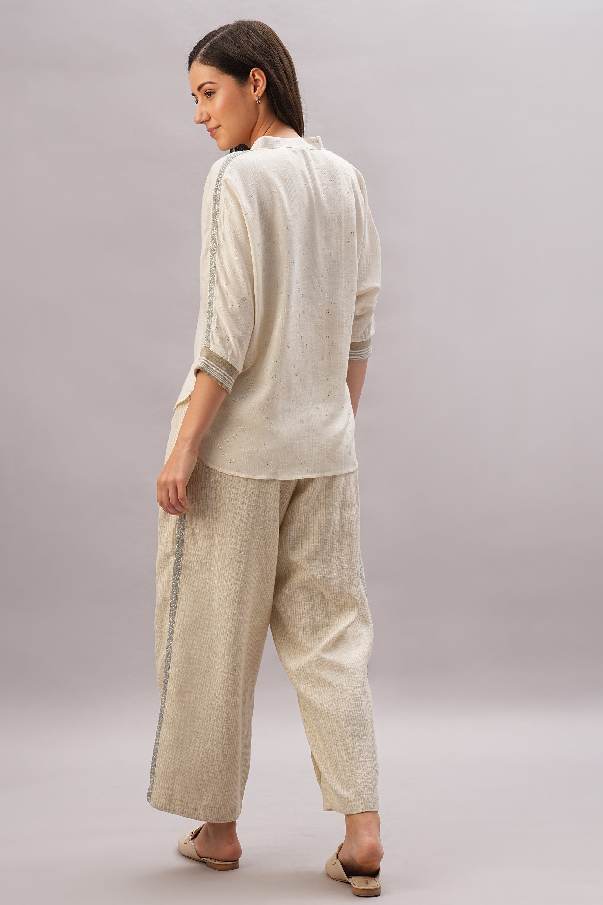 A woman stands barefoot, facing away, embodying effortless style and comfort in the Beige Linen set—featuring loose cream-colored pants and a long-sleeve top made from breathable linen.