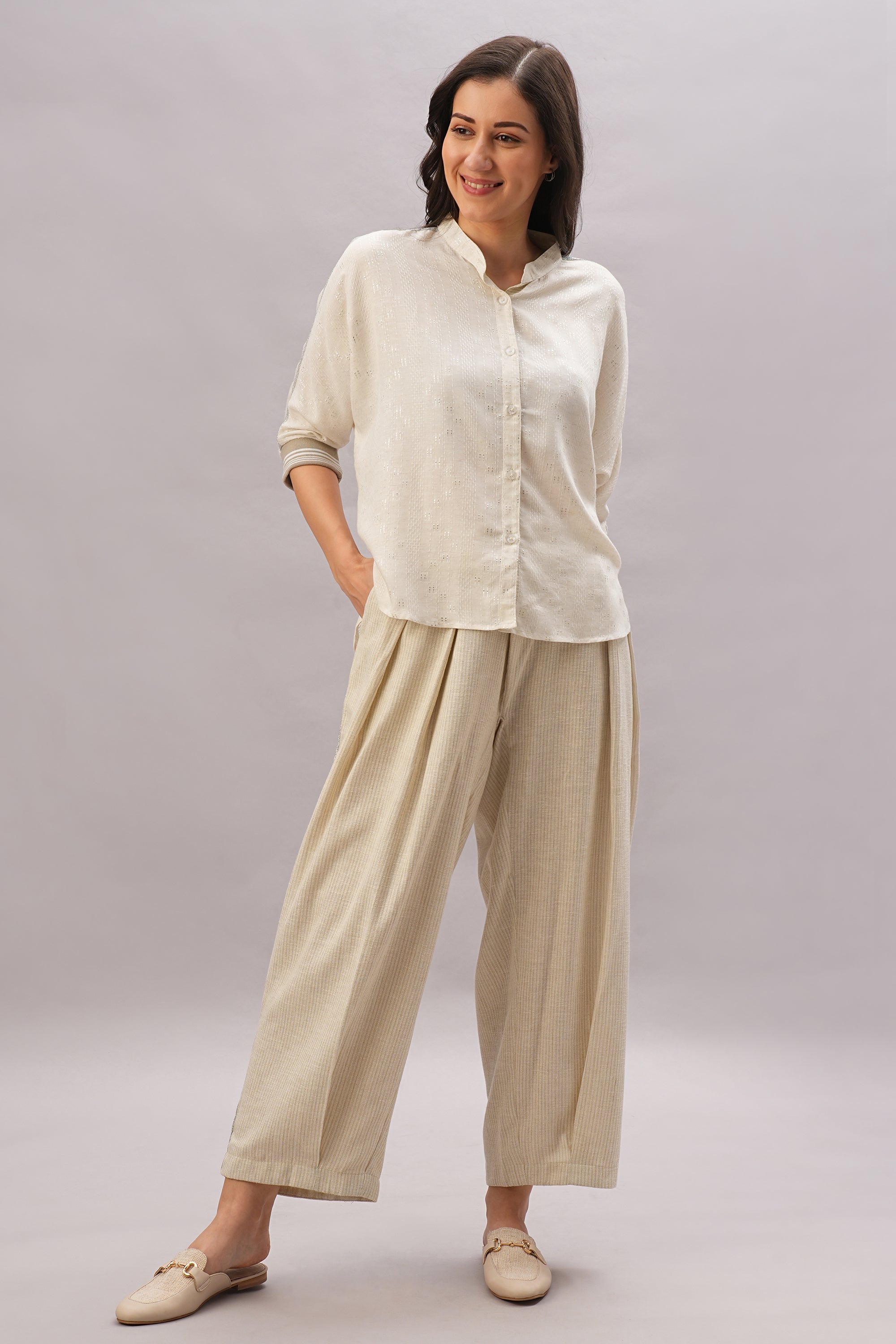 A woman smiles with hands in pockets, wearing the Beige Linen Set—a cream button-up shirt and loose beige pants—perfect for effortless style and breathability.