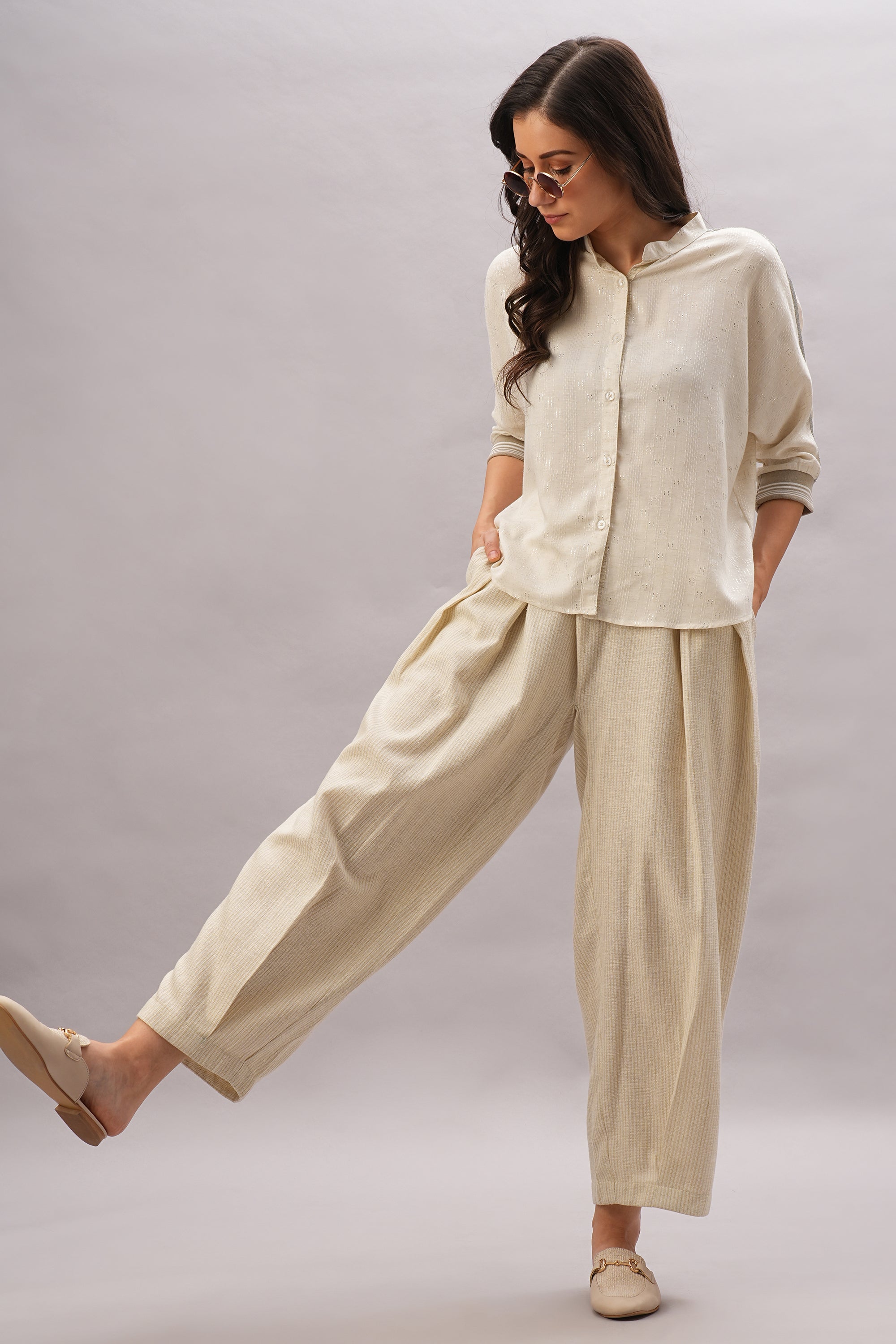 A woman models the Beige Linen set, featuring a loose shirt and wide pants in breathable linen, styled with sunglasses and flats as she stands confidently on one leg.