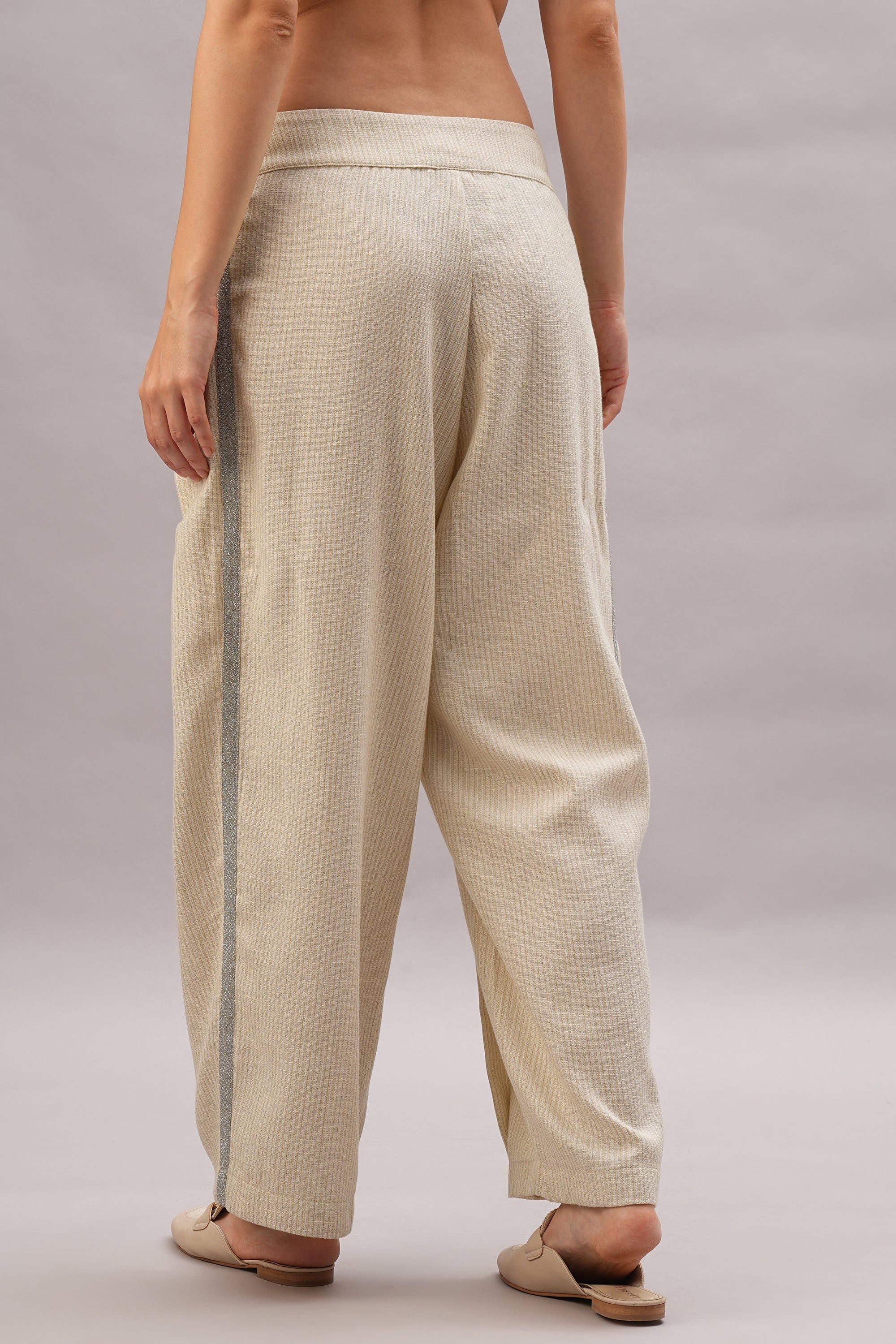 A person stands barefoot on a neutral background, showcasing the Beige Linen set—breathable, light-colored pants with a side stripe—effortless style captured from the back view.