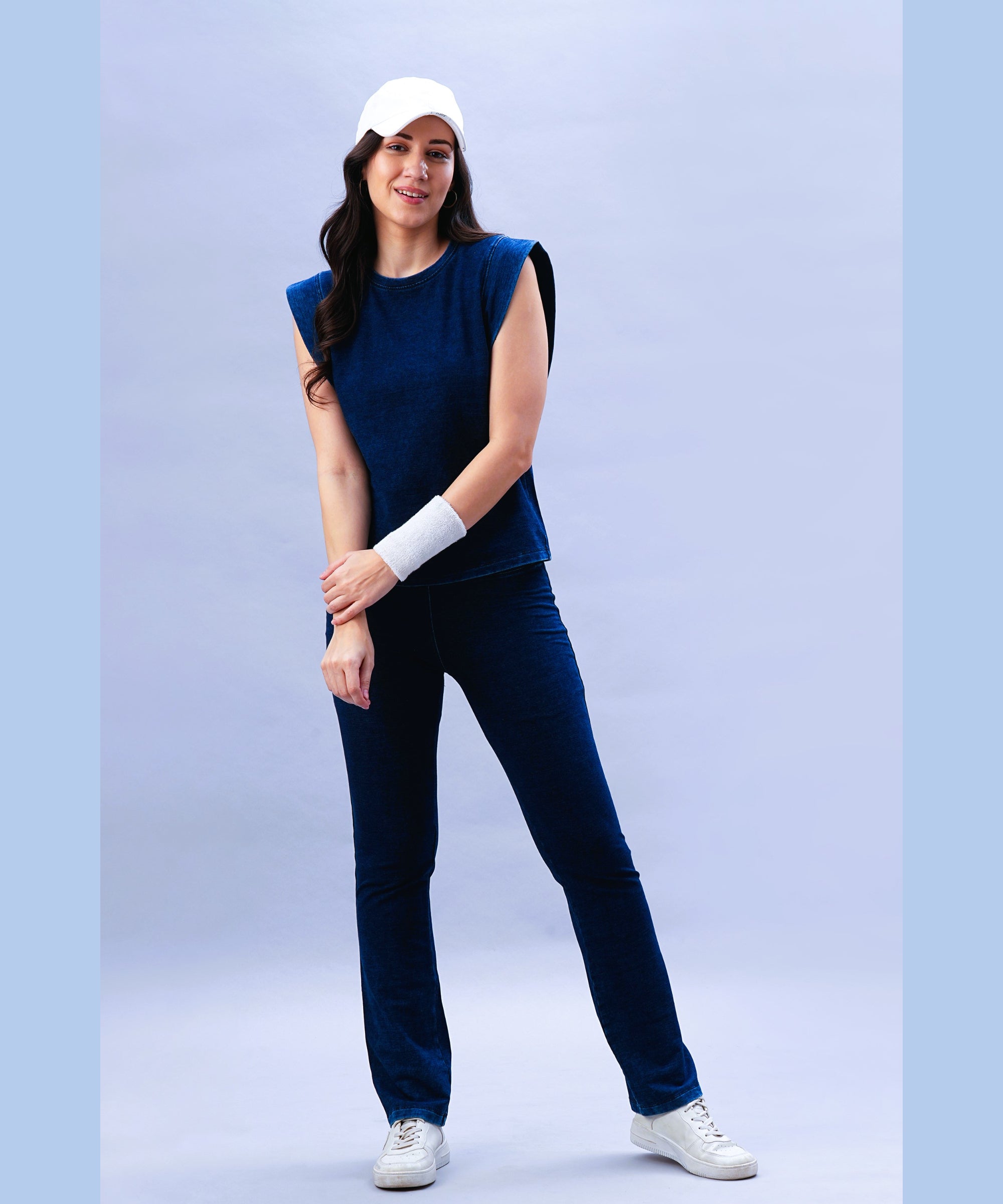 Woman in a blue sleeveless top, Blue Denim Wash Bootcut Leggings with a high-rise waistband, white sneakers, and a white cap, standing against a light blue background.