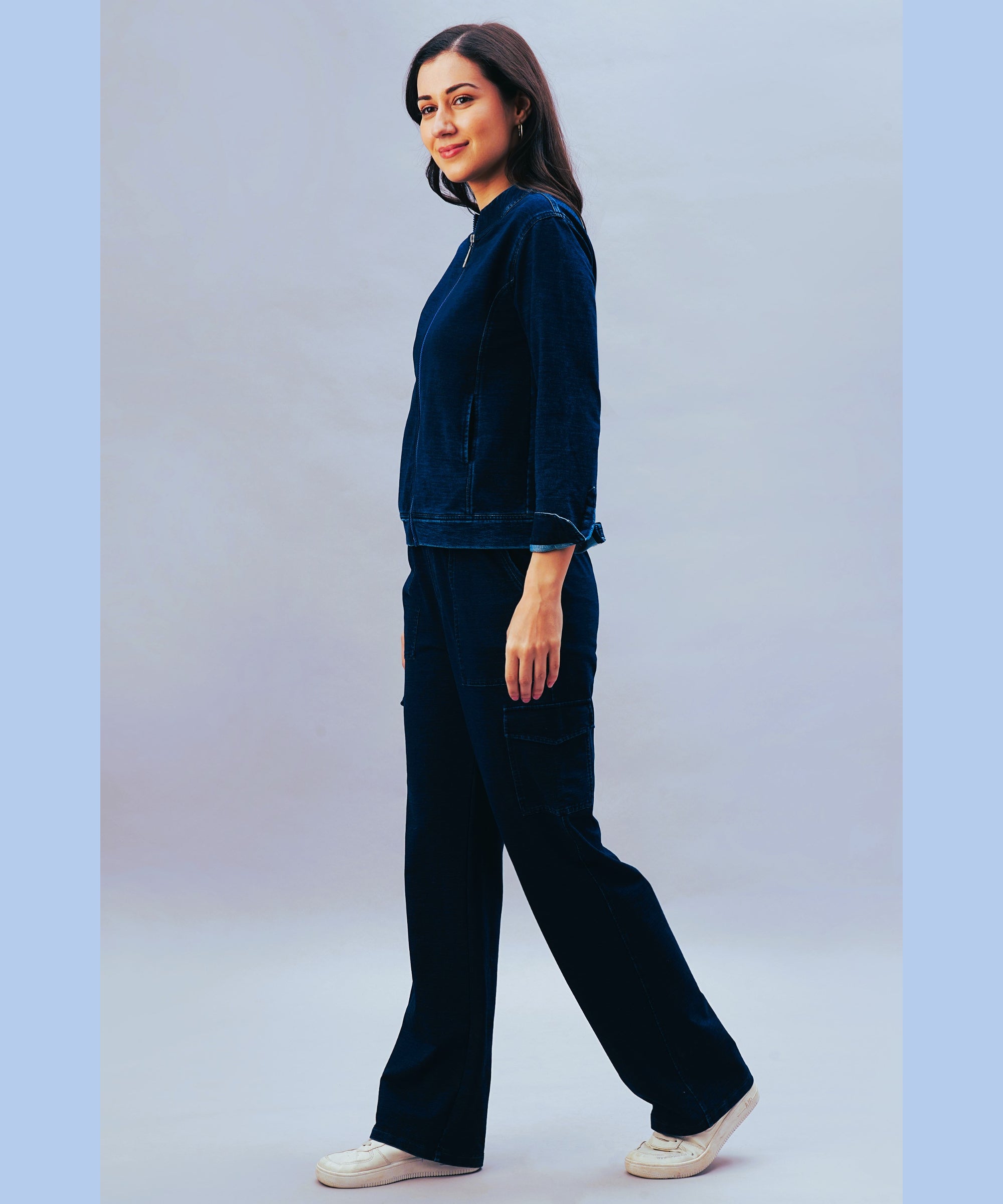 Woman in a dark blue outfit featuring the Blue Denim Wash Cargo Trackpant and white sneakers, standing sideways and smiling against a light blue background.
