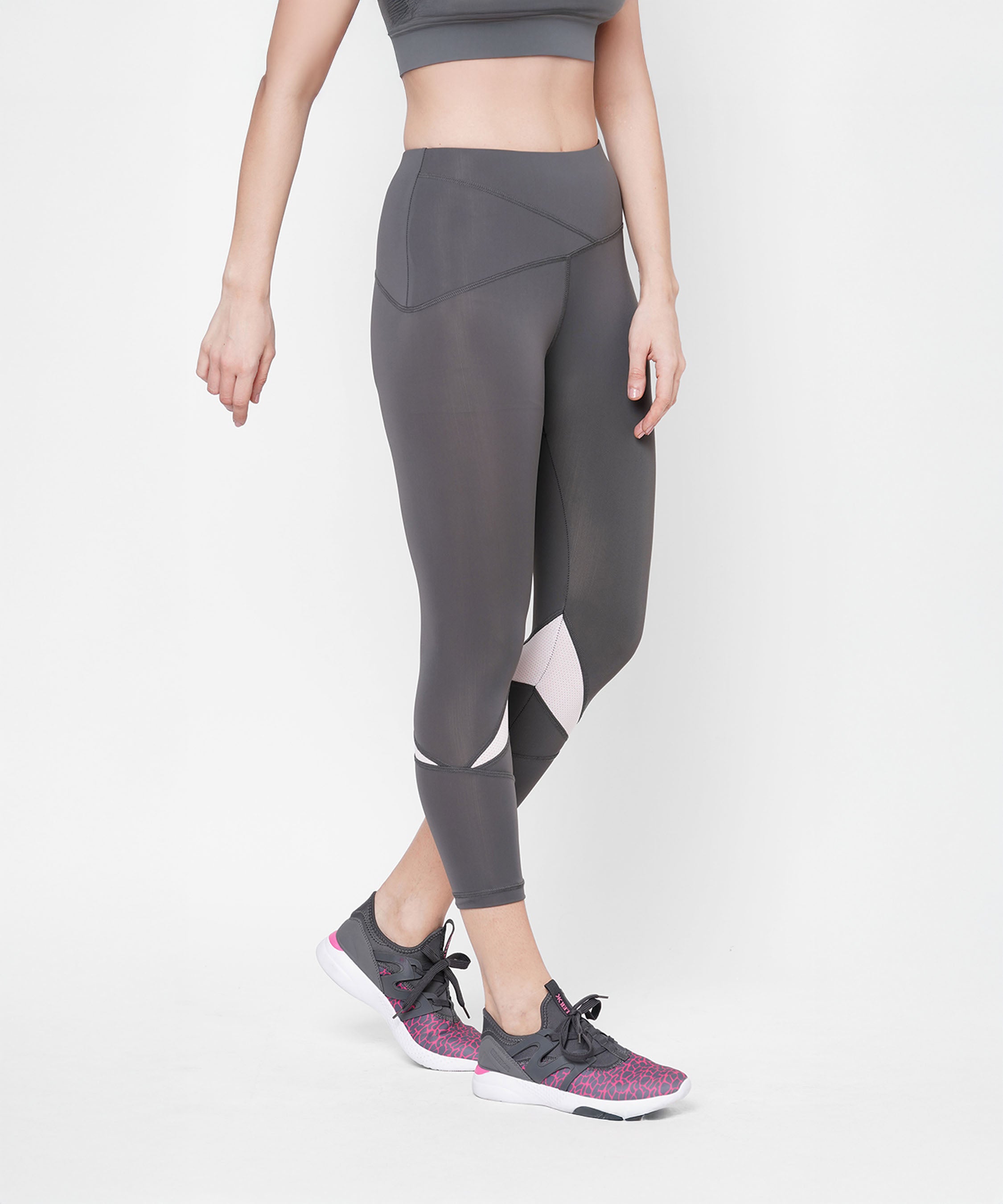 Woman wearing Classic Capri grey leggings with a thick waistband, paired with a gray sports bra and gray sneakers with pink accents, standing against a white background.