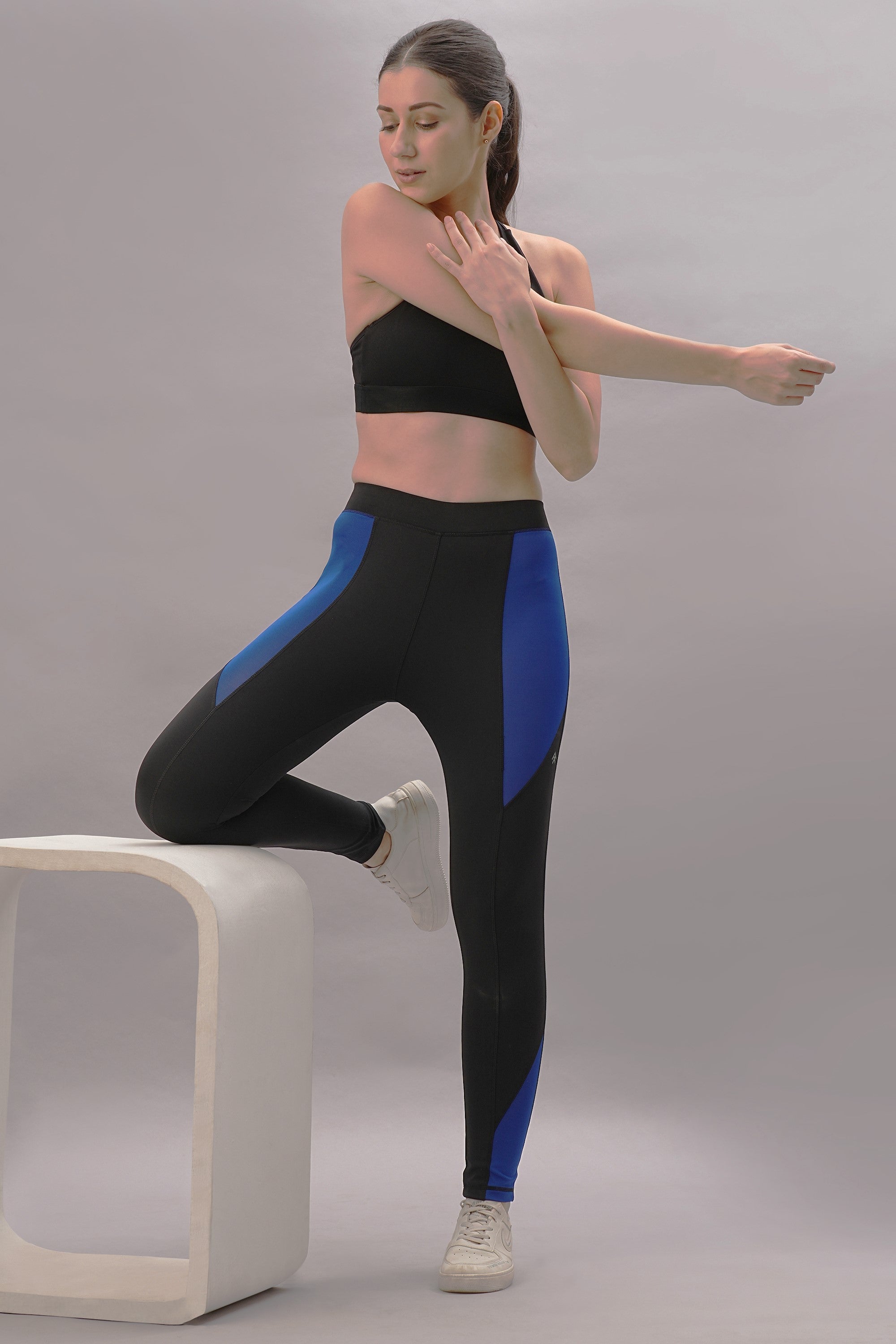 A woman in athletic wear stretches her arm with one foot on a white block, highlighting the Black and Blue Pants in a stylish, tailored fit against a gray background.