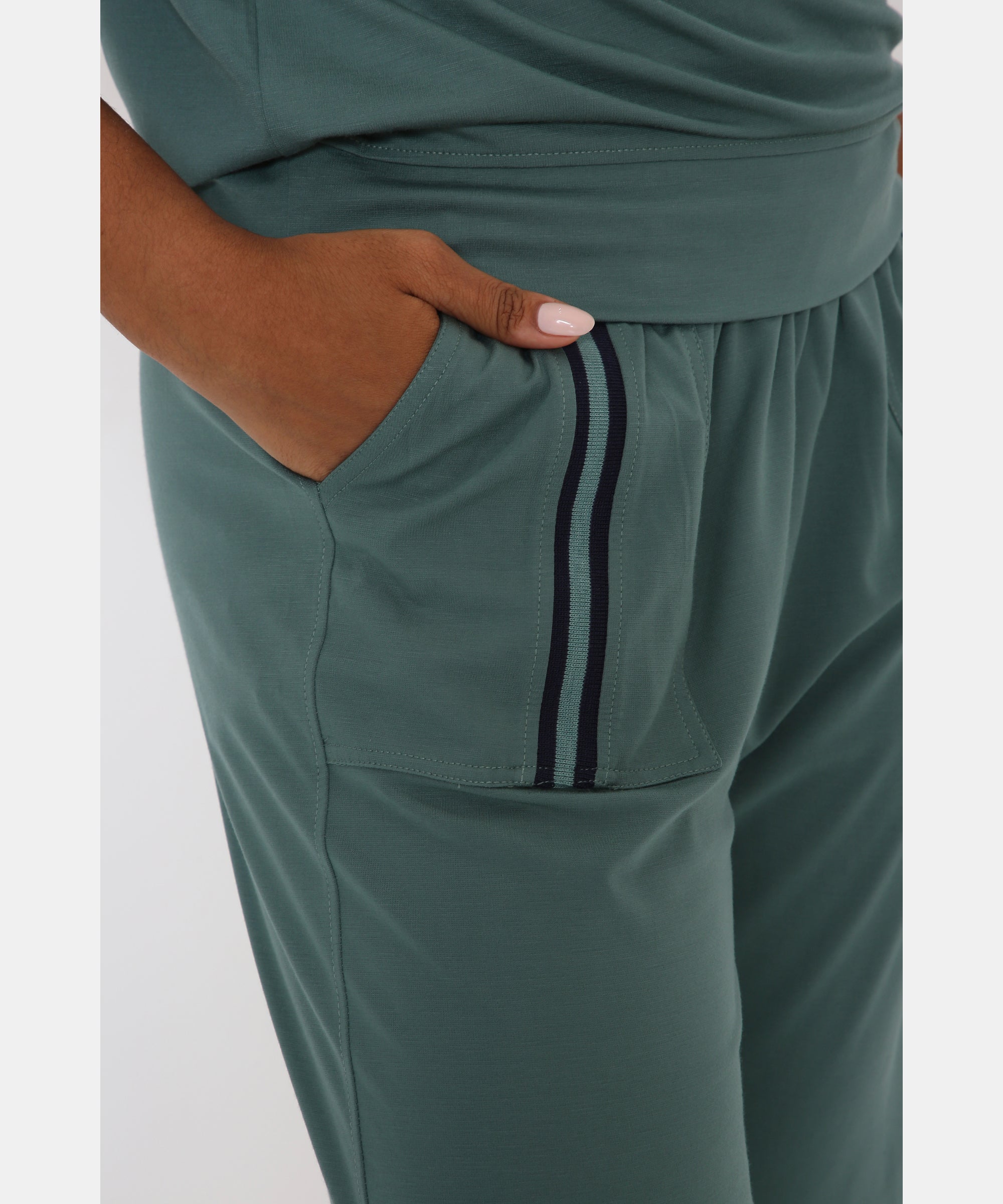 A person wearing the Sporty Sage Green Offshoulder Tracksuit Set, featuring jogger pants with dark stripes and a hand in the pocket.