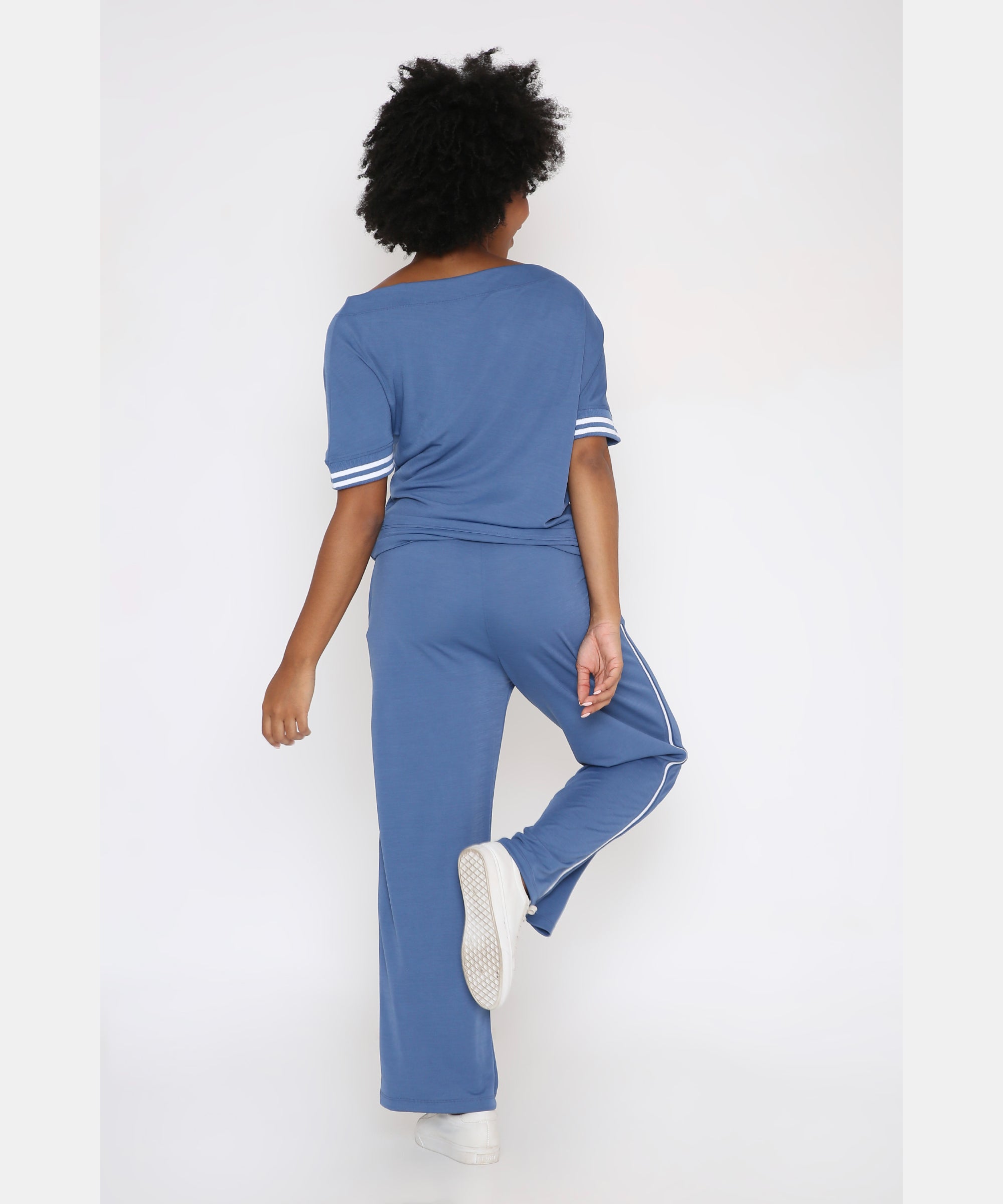 Model with curly hair wears the Sporty Atlantic Blue Off-Shoulder Tracksuit Set, posing with one leg raised and facing away from the camera against a white background.