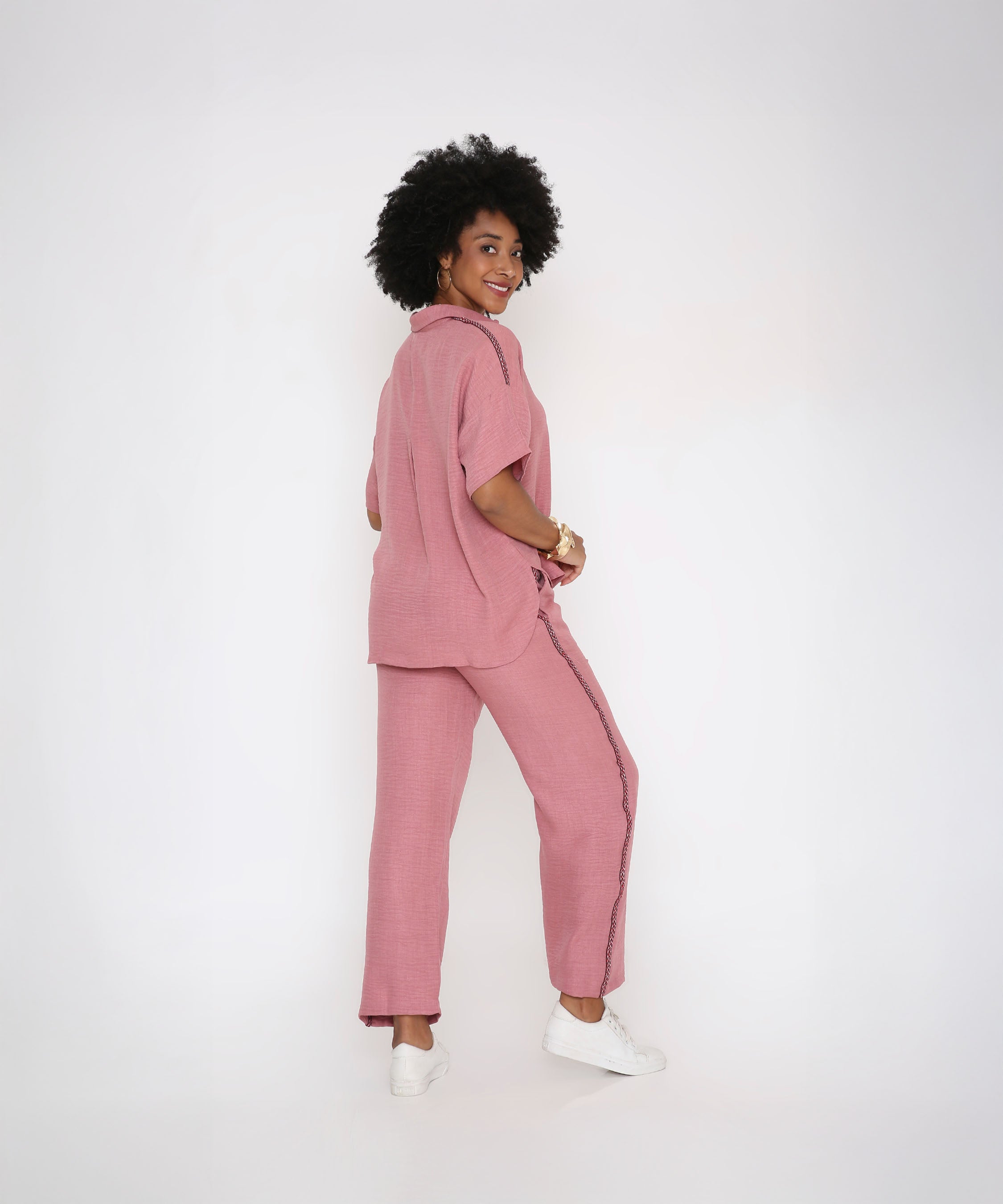 A woman smiles and looks over her shoulder in the Vintage Pink Loose Shirt and Pant Cord Set, made from soft viscose cotton, styled with classic white sneakers against a white background.