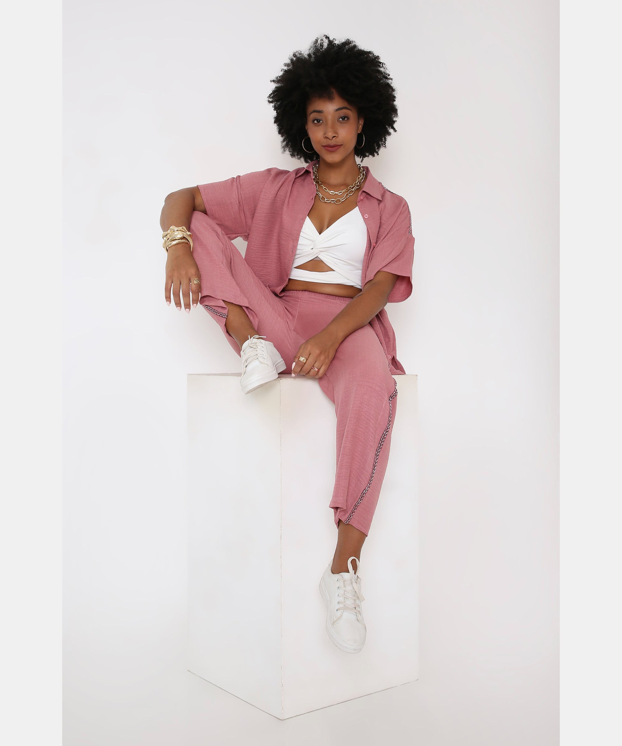 A woman with curly hair wears the Vintage Pink Loose Shirt and Pant Cord Set with a white top, seated on a white cube against a plain background.