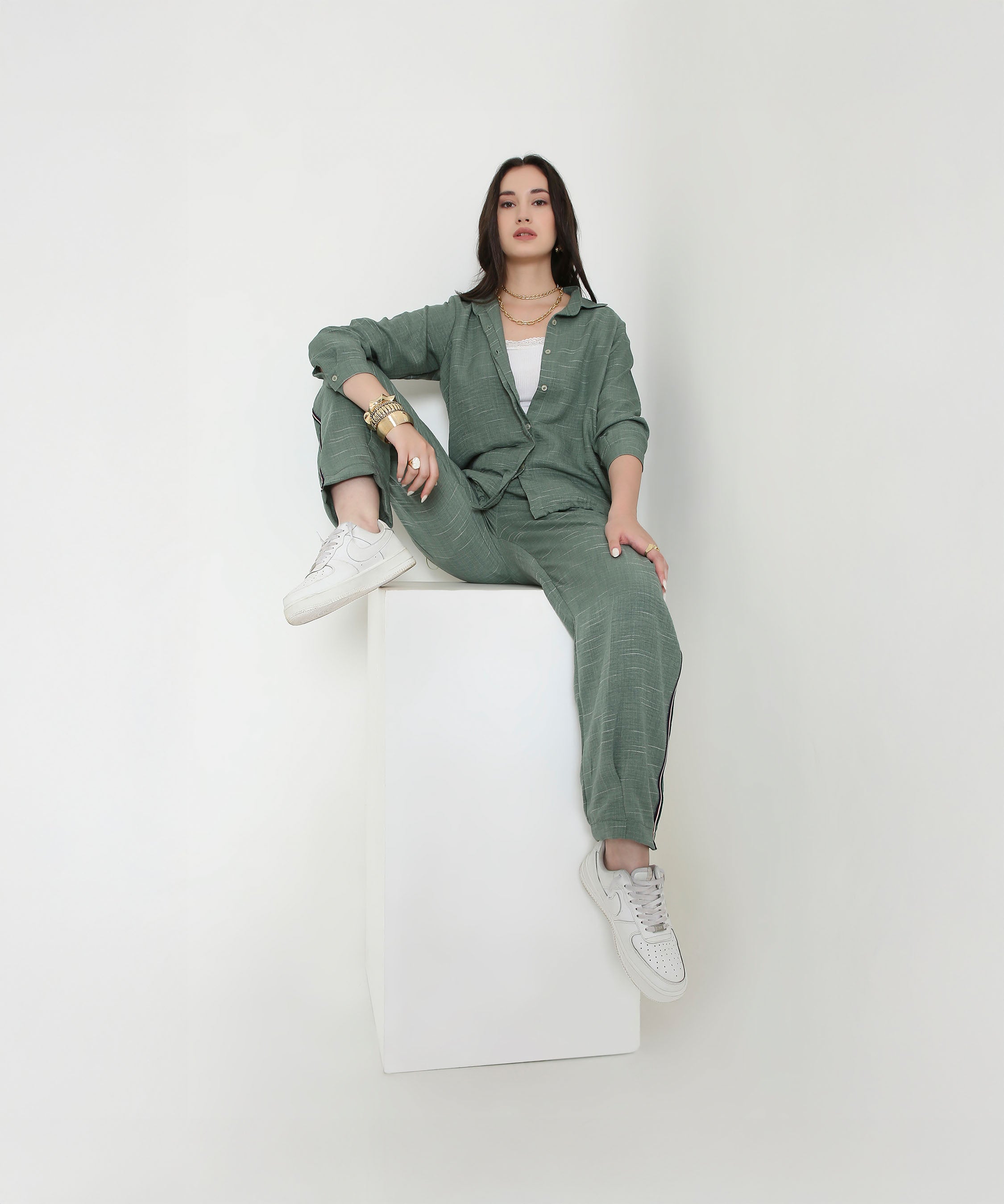 A woman in the Moss Green Breezy Cord Set and white sneakers sits confidently on a white cube against a plain background, highlighting the relaxed fit and modern style of this loungewear.
