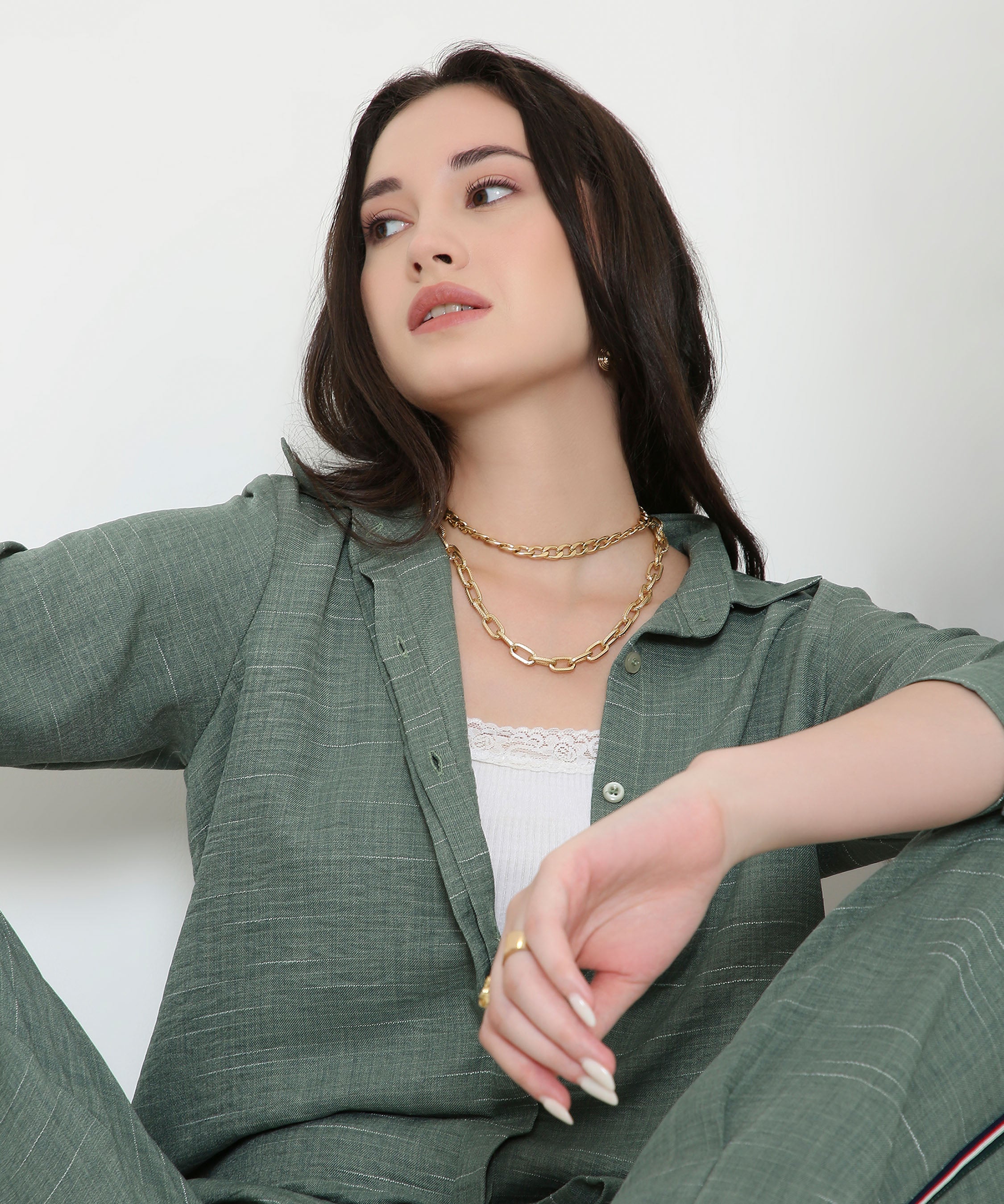 A woman sits looking to the side, styled in the Moss Green Breezy Cord Set with layered necklaces over a white top.