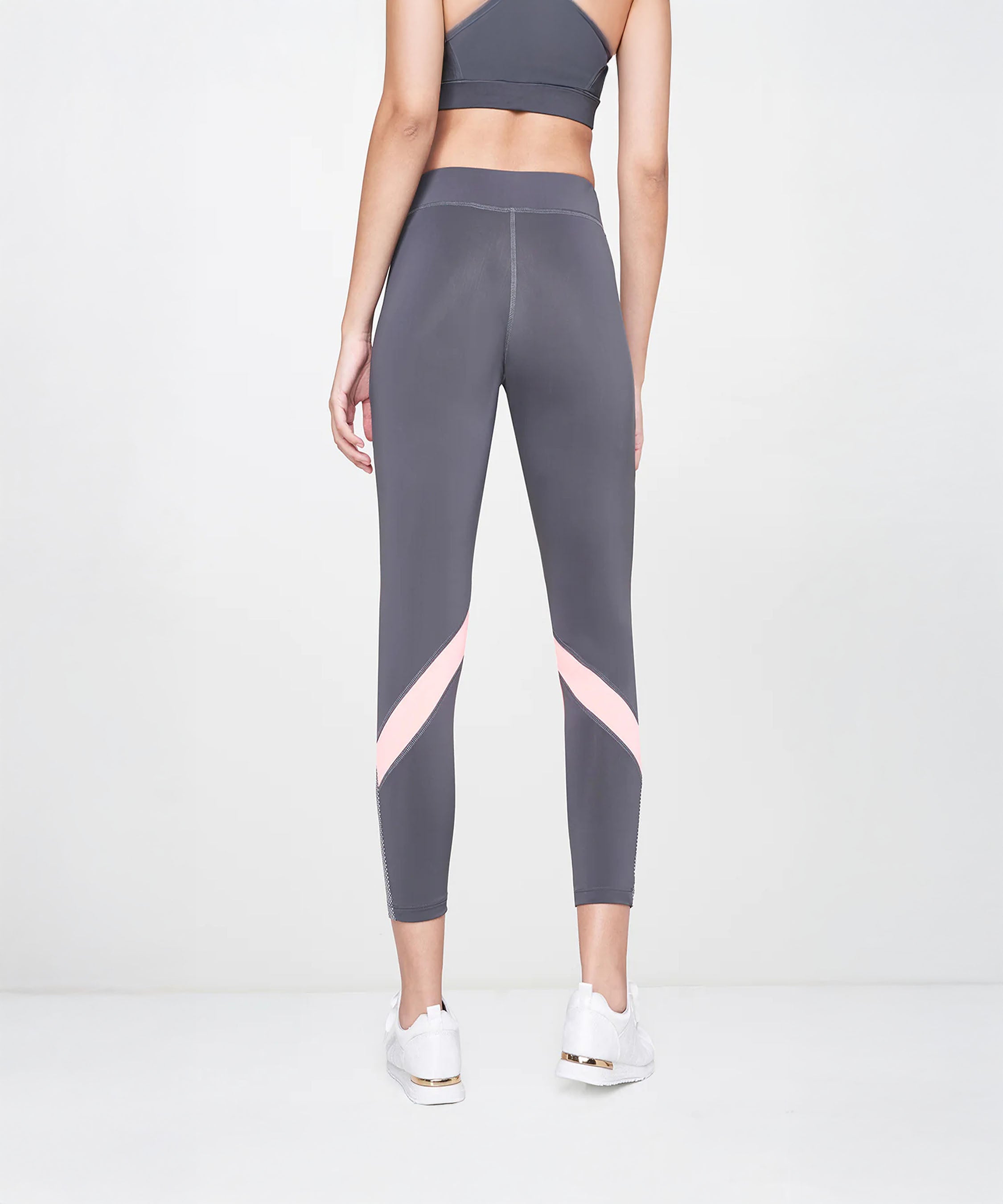 A woman stands facing away on a plain light background, wearing Grey Leggings featuring pink stripes and color-block panels.