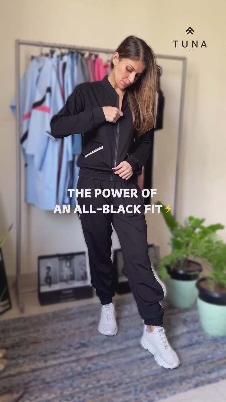 Load video: Woman in a black tracksuit, white sneakers, and a black cap standing against a light gray background.